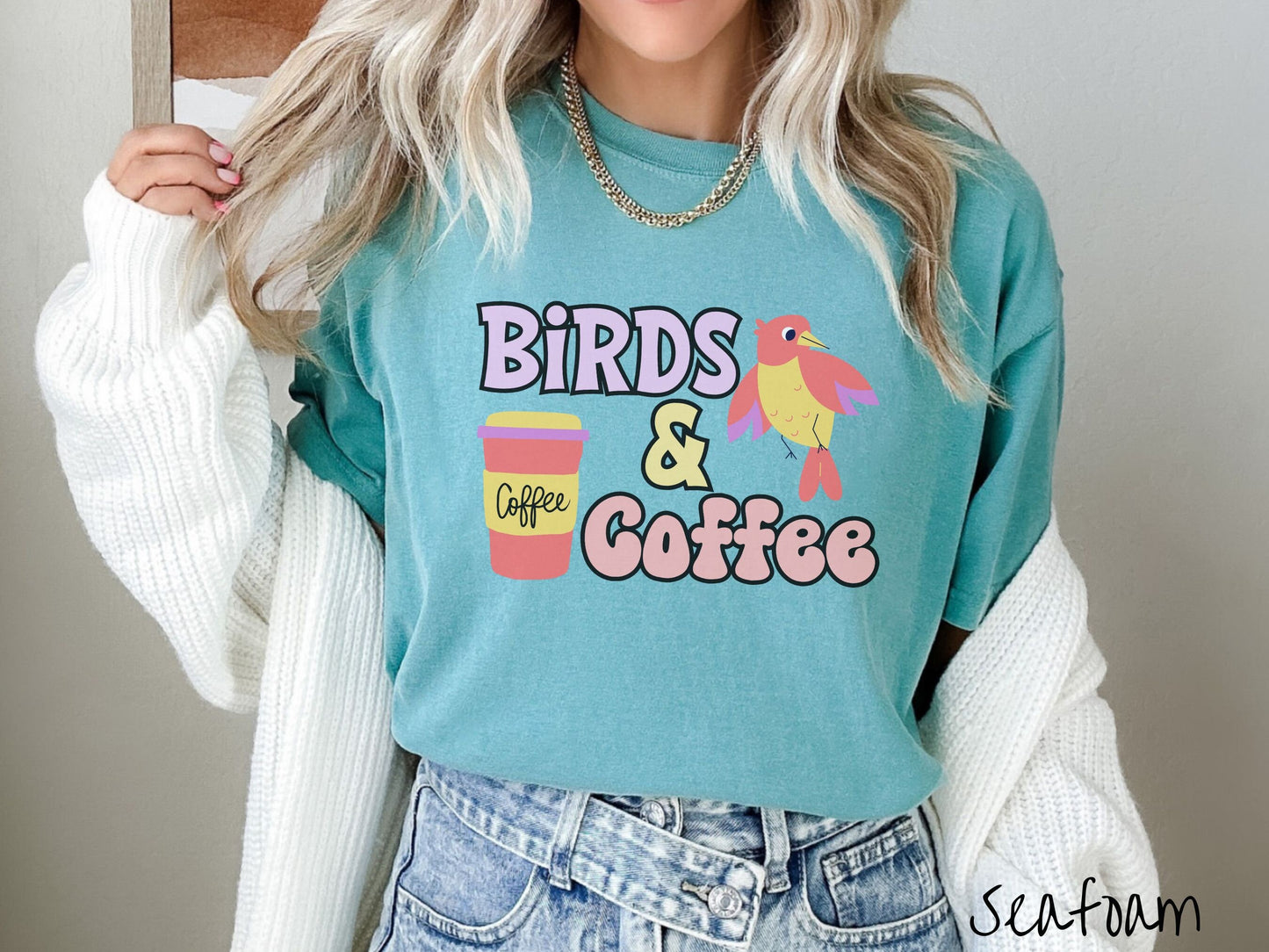 A woman wearing a cute, vintage seafoam colored Comfort Colors shirt with the text Birds & Coffee in purple and light pink font. Next to the text is a red, yellow, and purple smiling bird in the air and a red and yellow to-go coffee cup.