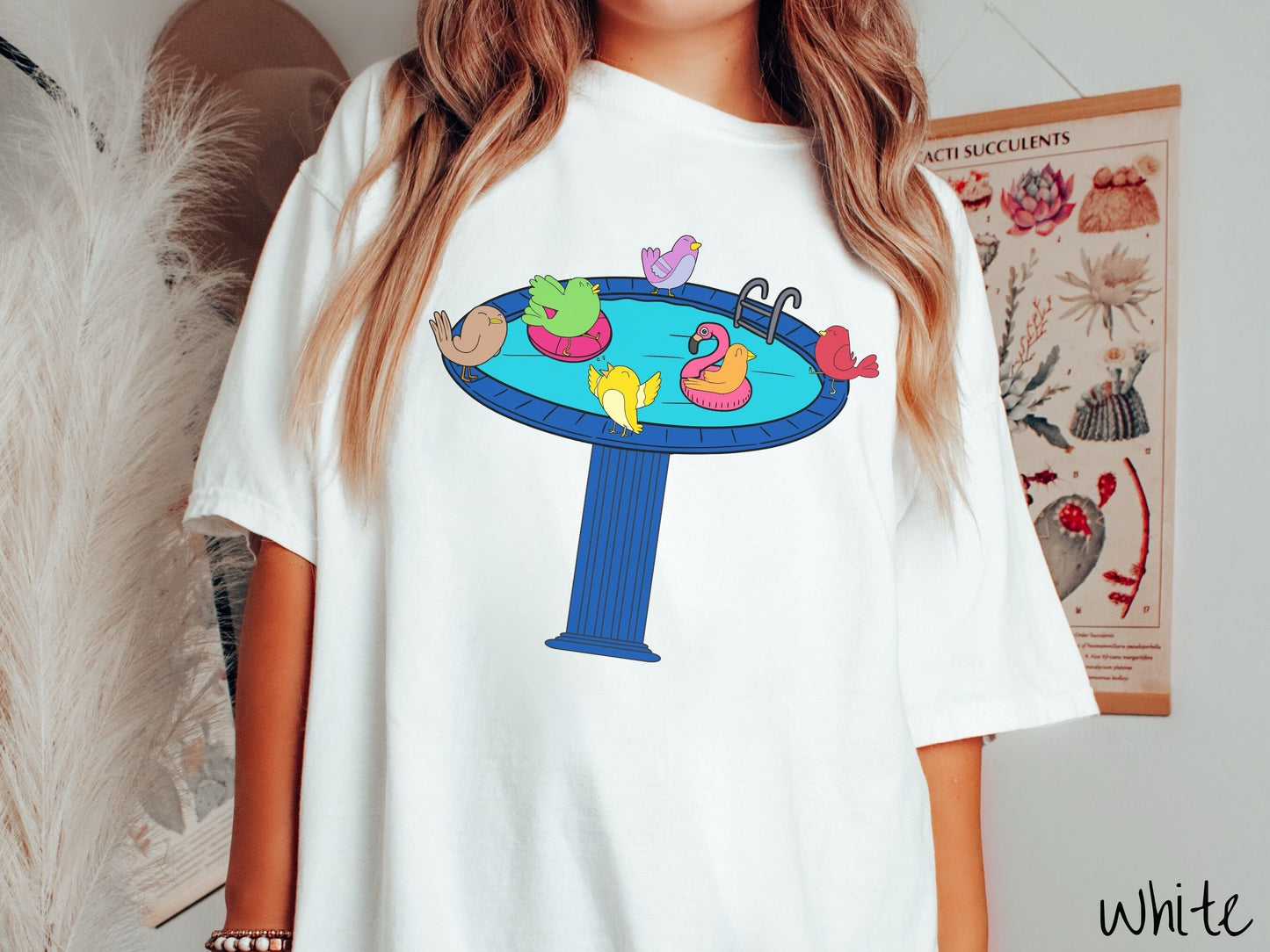 A woman wearing a cute, vintage white colored Comfort Colors shirt with a blue birdbath with a pool ladder filled with crystal clear blue water. In the water are colorful birds floating on colorful pool inflatables and enjoying the sun.