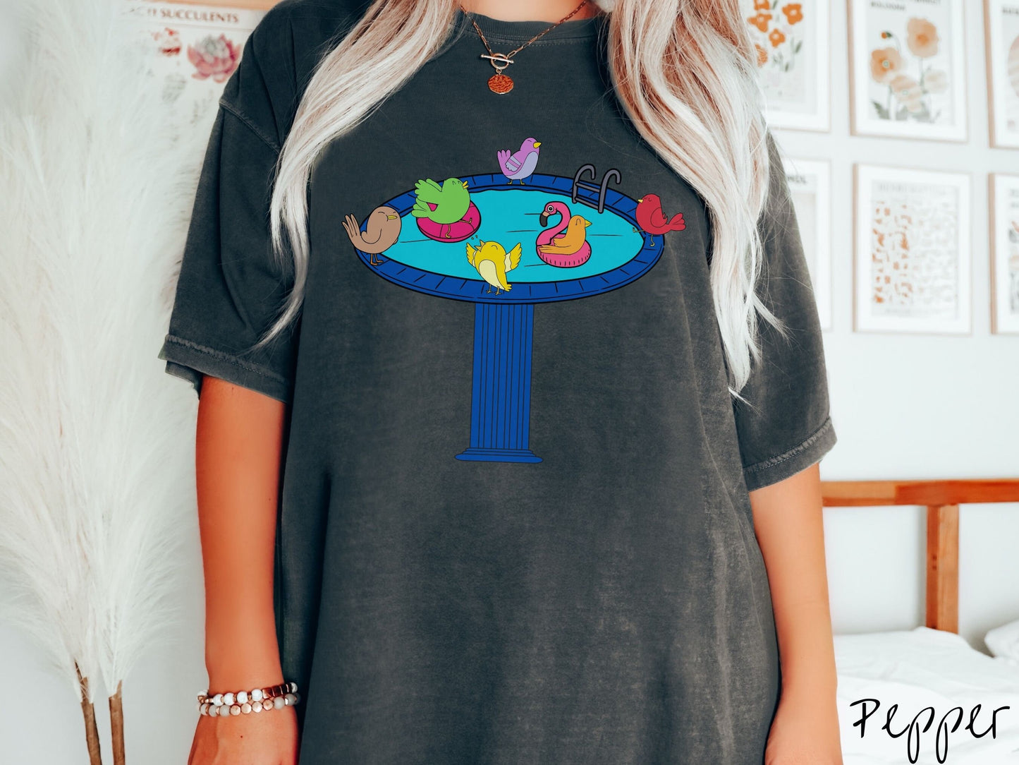 A woman wearing a cute, vintage pepper colored Comfort Colors shirt with a blue birdbath with a pool ladder filled with crystal clear blue water. In the water are colorful birds floating on colorful pool inflatables and enjoying the sun.