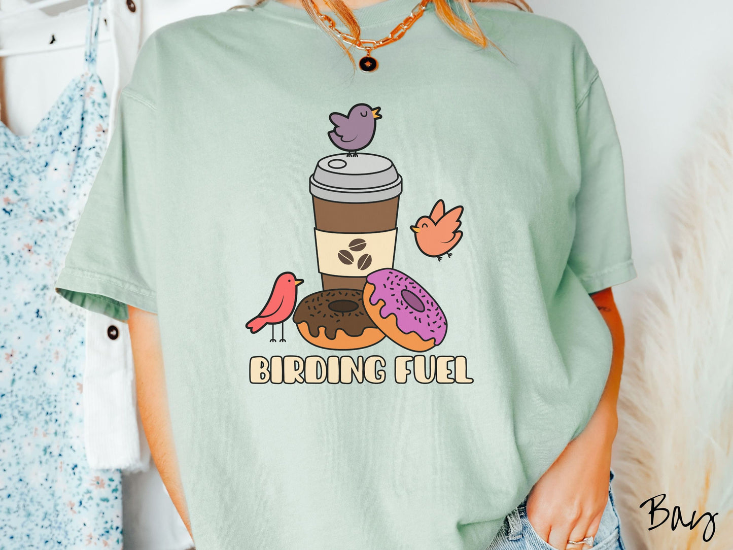 A woman wearing a cute, vintage bay colored Comfort Colors shirt with a brown to-go coffee cup next to a chocolate frosted donut and pink frosted donut. There are three orange, red, and purple birds sitting on the coffee cup and flying around.