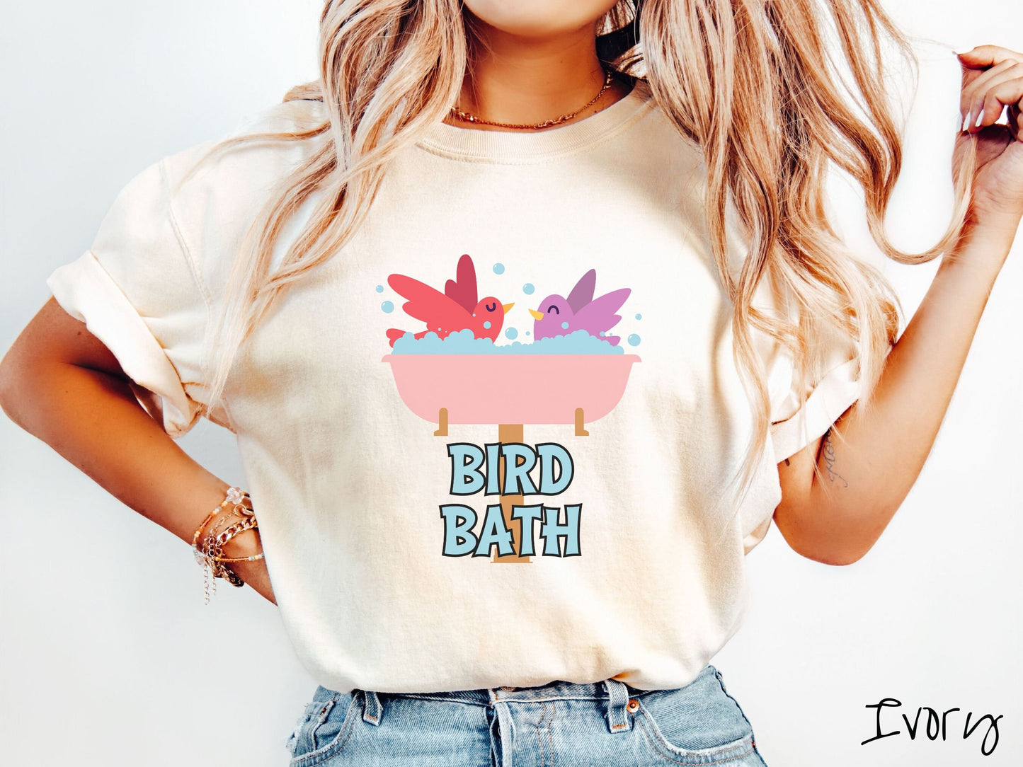 A woman wearing a cute, vintage ivory colored Comfort Colors shirt with a pink bathtub with a red bird and purple bird enjoying a bubble bath together. Below the bathtub are the words Bird Bath in light blue, uppercase font.