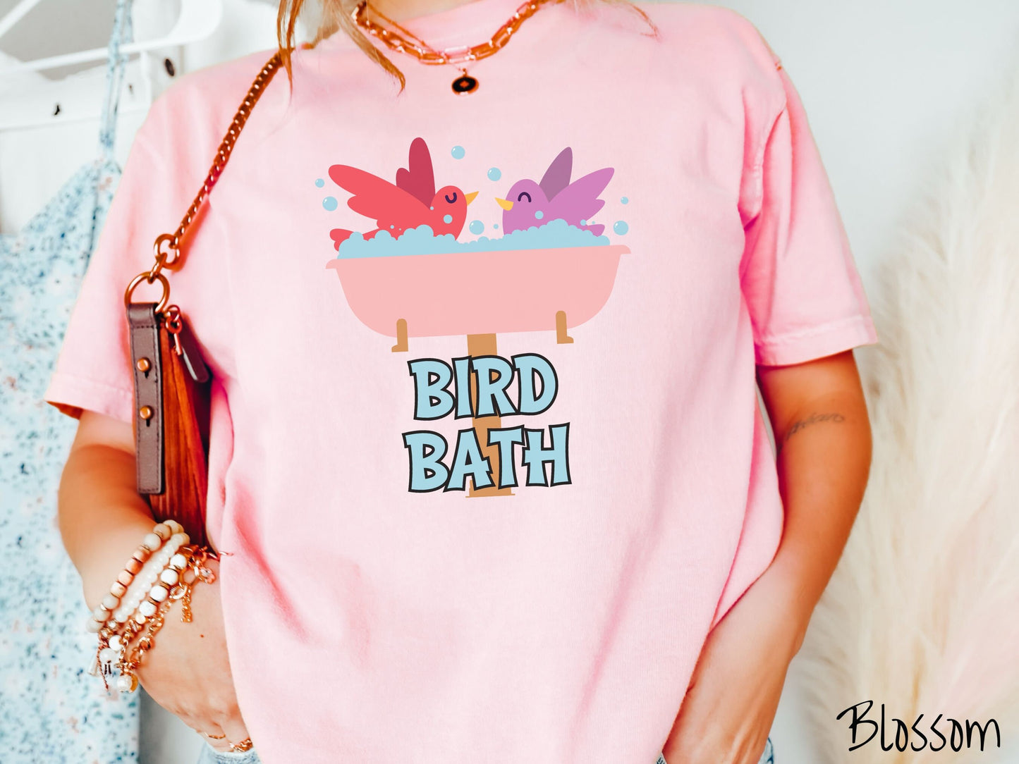 A woman wearing a cute, vintage blossom colored Comfort Colors shirt with a pink bathtub with a red bird and purple bird enjoying a bubble bath together. Below the bathtub are the words Bird Bath in light blue, uppercase font.