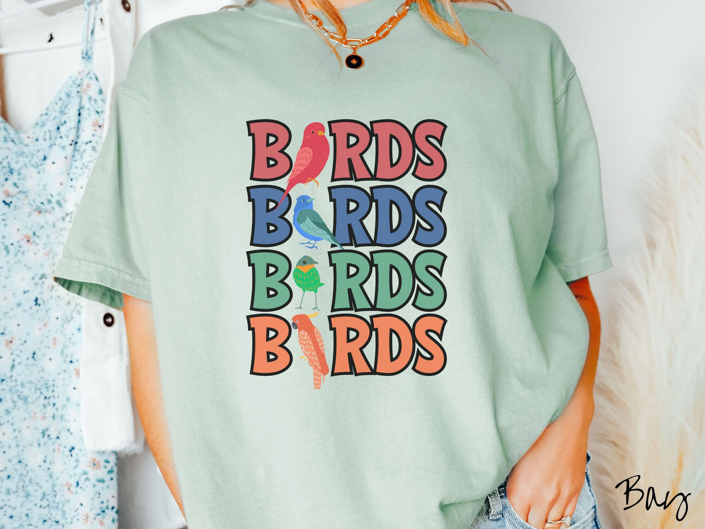 A woman wearing a cute, vintage bay colored Comfort Colors t-shirt with the word BIRDS listed four times vertically in red, blue, green, and orange uppercase font. The Is in BIRDS are different kinds of colorful birds.