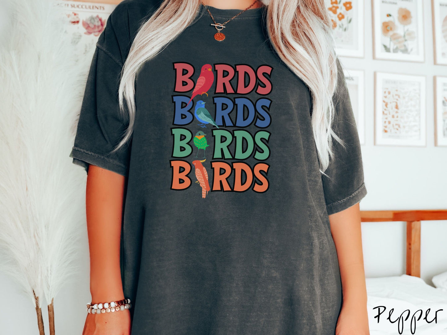 A woman wearing a cute, vintage pepper colored Comfort Colors t-shirt with the word BIRDS listed four times vertically in red, blue, green, and orange uppercase font. The Is in BIRDS are different kinds of colorful birds.