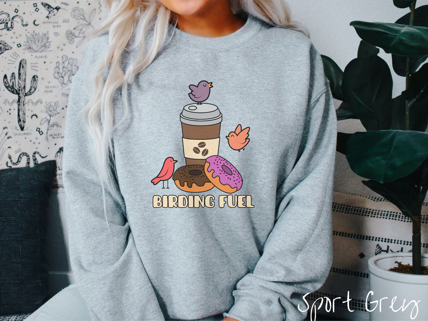 A woman wearing a cute, vintage sport grey colored comfy sweatshirt with a brown to-go coffee cup next to a chocolate frosted donut and pink frosted donut. There are three orange, red, and purple birds sitting on the coffee cup and flying around.