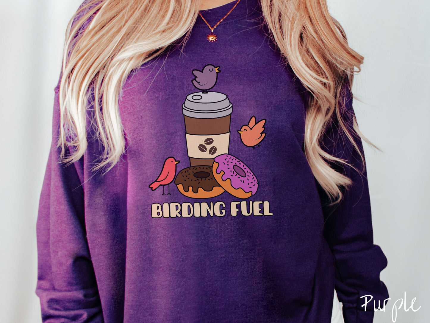 A woman wearing a cute, vintage purple colored comfy sweatshirt with a brown to-go coffee cup next to a chocolate frosted donut and pink frosted donut. There are three orange, red, and purple birds sitting on the coffee cup and flying around.