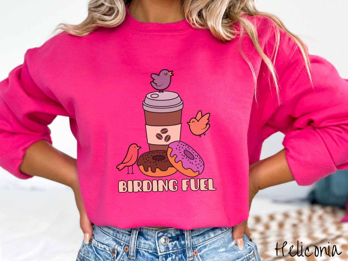 A woman wearing a cute, vintage heliconia colored comfy sweatshirt with a brown to-go coffee cup next to a chocolate frosted donut and pink frosted donut. There are three orange, red, and purple birds sitting on the coffee cup and flying around.