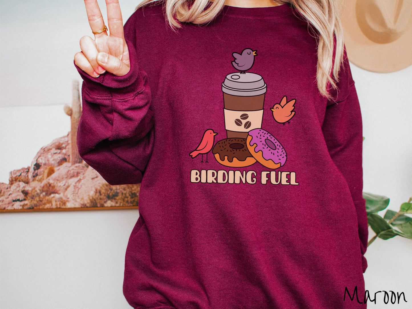 A woman wearing a cute, vintage maroon colored comfy sweatshirt with a brown to-go coffee cup next to a chocolate frosted donut and pink frosted donut. There are three orange, red, and purple birds sitting on the coffee cup and flying around.
