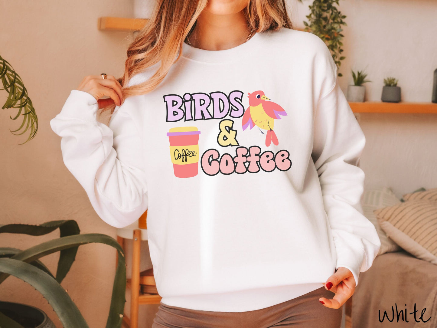 A woman wearing a cute, vintage white colored comfy sweatshirt with the text Birds & Coffee in purple and light pink font. Next to the text is a red, yellow, and purple smiling bird in the air and a red and yellow to-go coffee cup.
