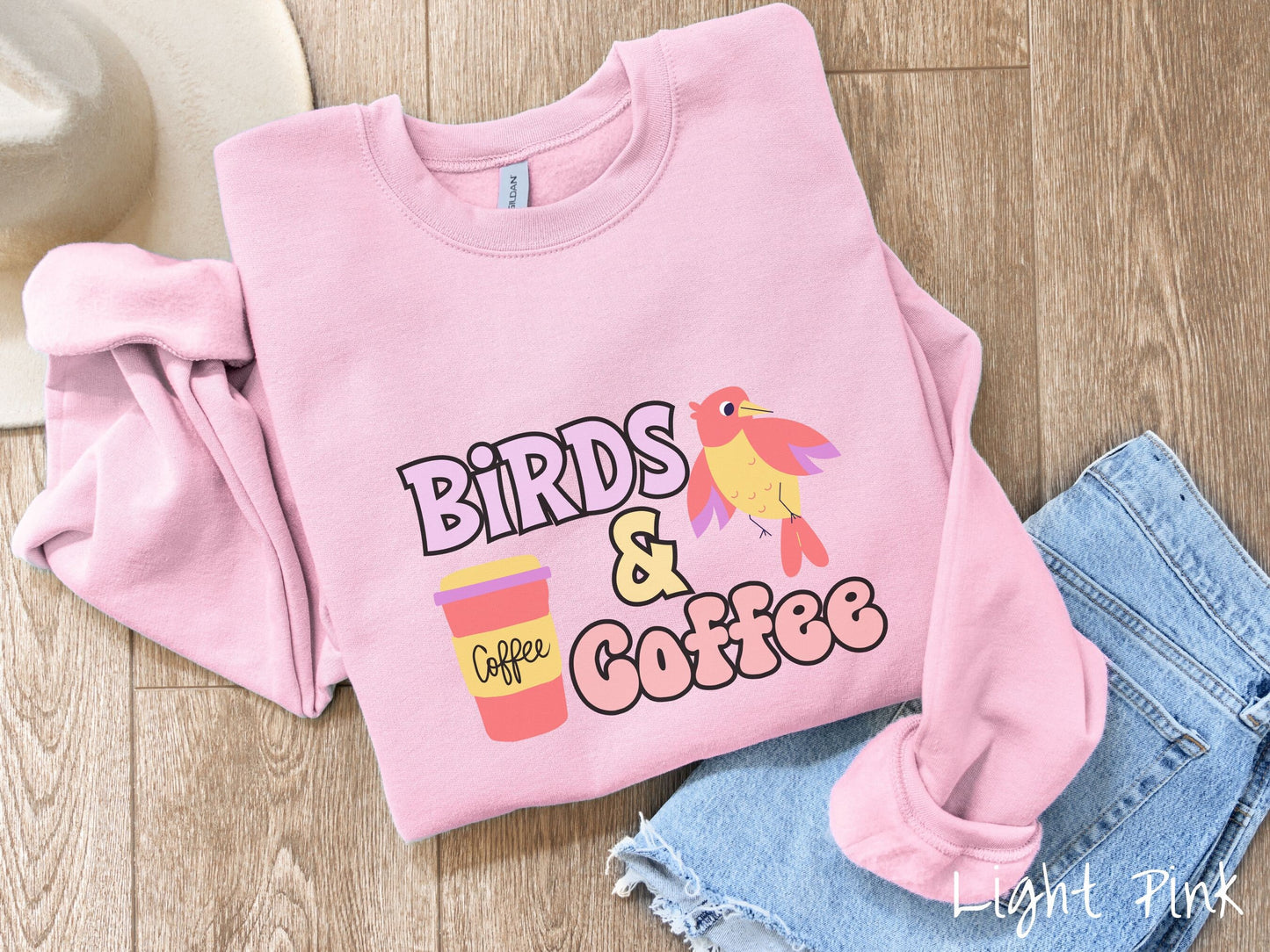 A cute, vintage light pink colored comfy sweatshirt with the text Birds & Coffee in purple and light pink font. Next to the text is a red, yellow, and purple smiling bird in the air and a red and yellow to-go coffee cup.
