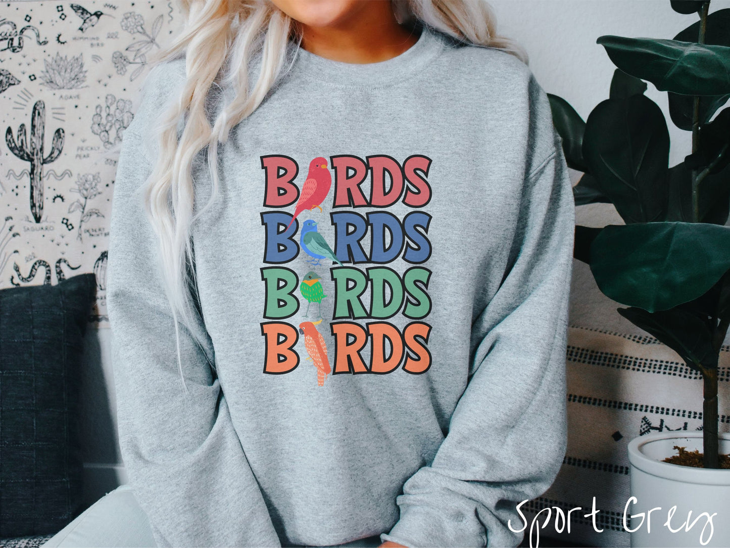 A woman wearing a cute, vintage sport grey colored comfy sweatshirt with the word BIRDS listed four times vertically in red, blue, green, and orange uppercase font. The Is in BIRDS are different kinds of colorful birds.