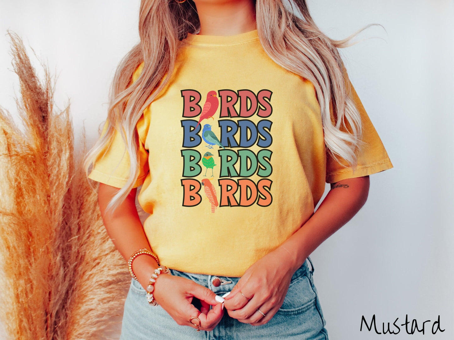 A woman wearing a cute, vintage mustard colored Comfort Colors t-shirt with the word BIRDS listed four times vertically in red, blue, green, and orange uppercase font. The Is in BIRDS are different kinds of colorful birds.