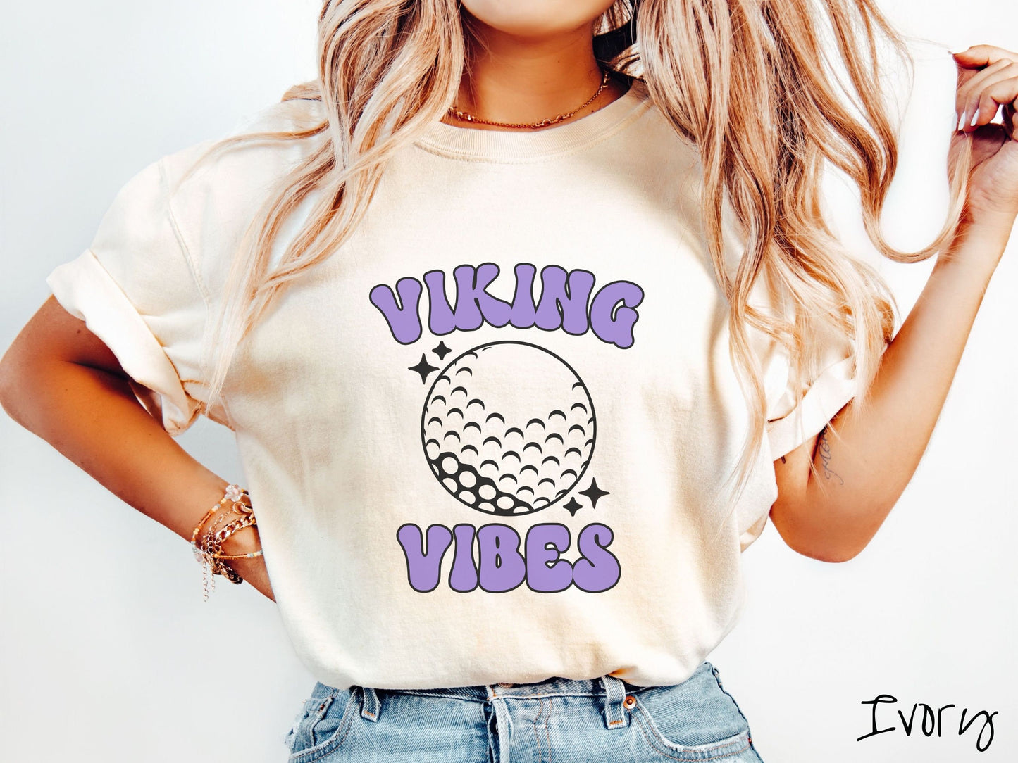 A woman wearing a cute ivory colored t-shirt with purple text saying Viking Vibes. There is a golf ball outline with black twinkling stars around it between the text.