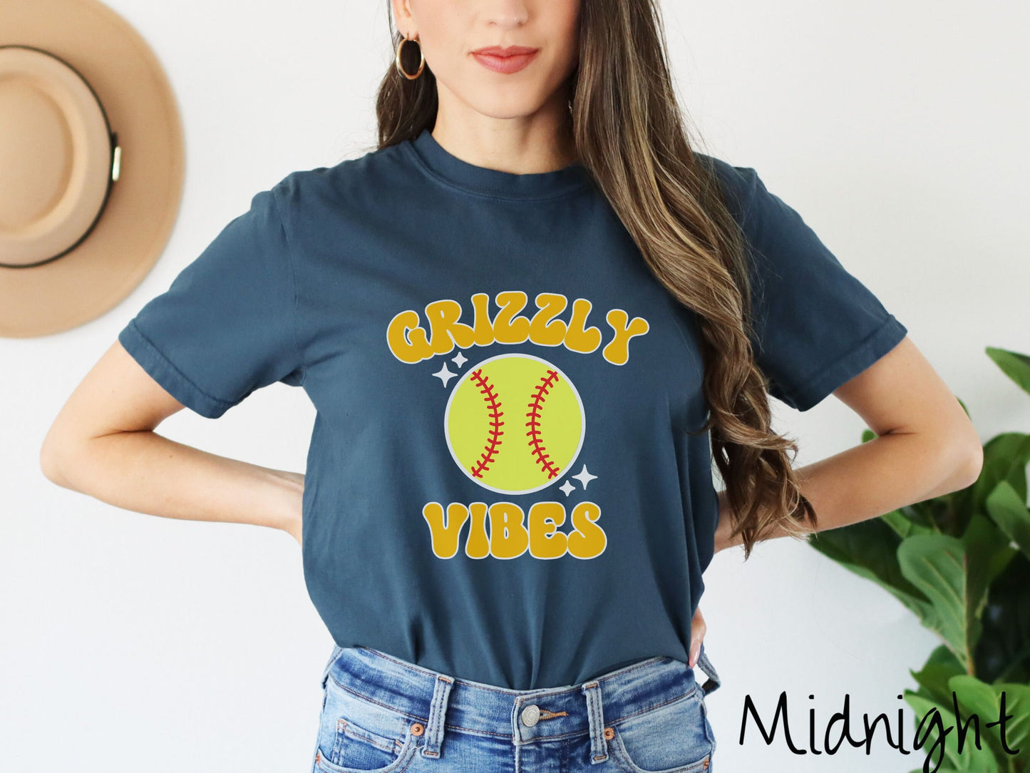 A woman wearing a cute midnight colored shirt with orange text saying Grizzly Vibes. There is a yellow baseball with red stitching with black twinkling stars around it between the text.
