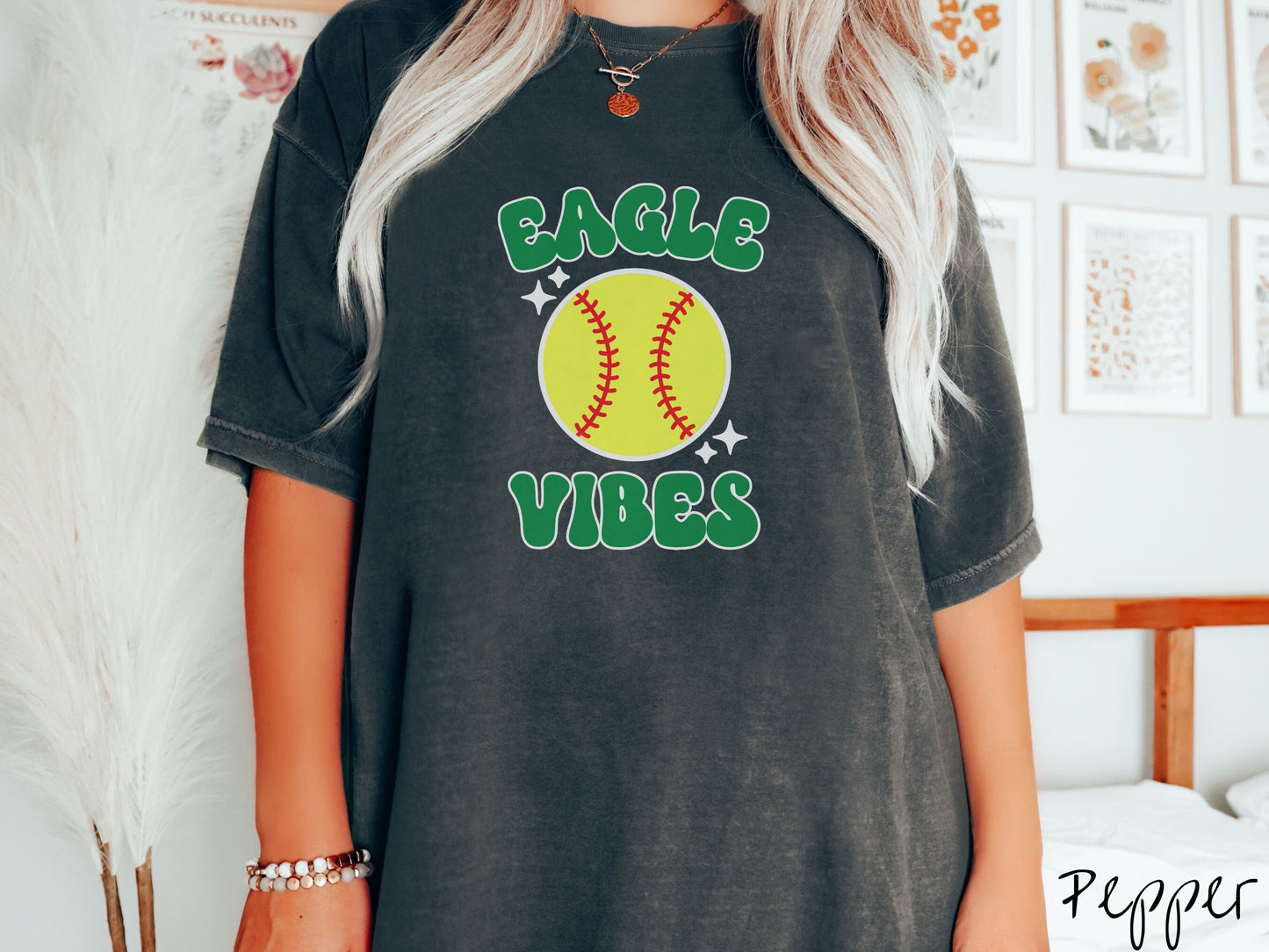 A woman wearing a cute pepper colored shirt with green text saying Eagle Vibes. There is a yellow baseball with red stitching with black twinkling stars around it between the text.