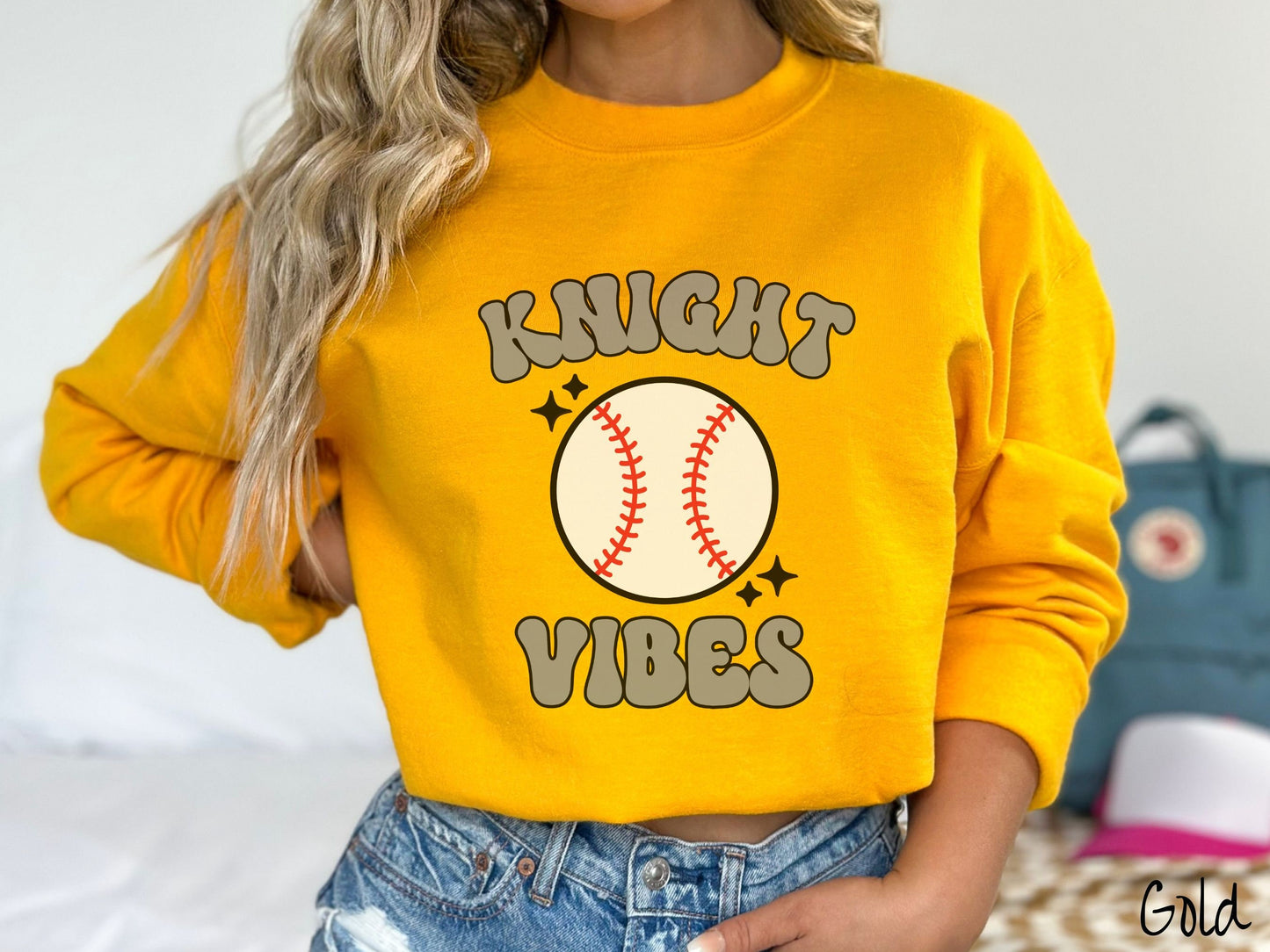 A woman wearing a cute gold colored sweatshirt with gray text saying Panther Vibes. There is a white baseball with red stitching between the text.