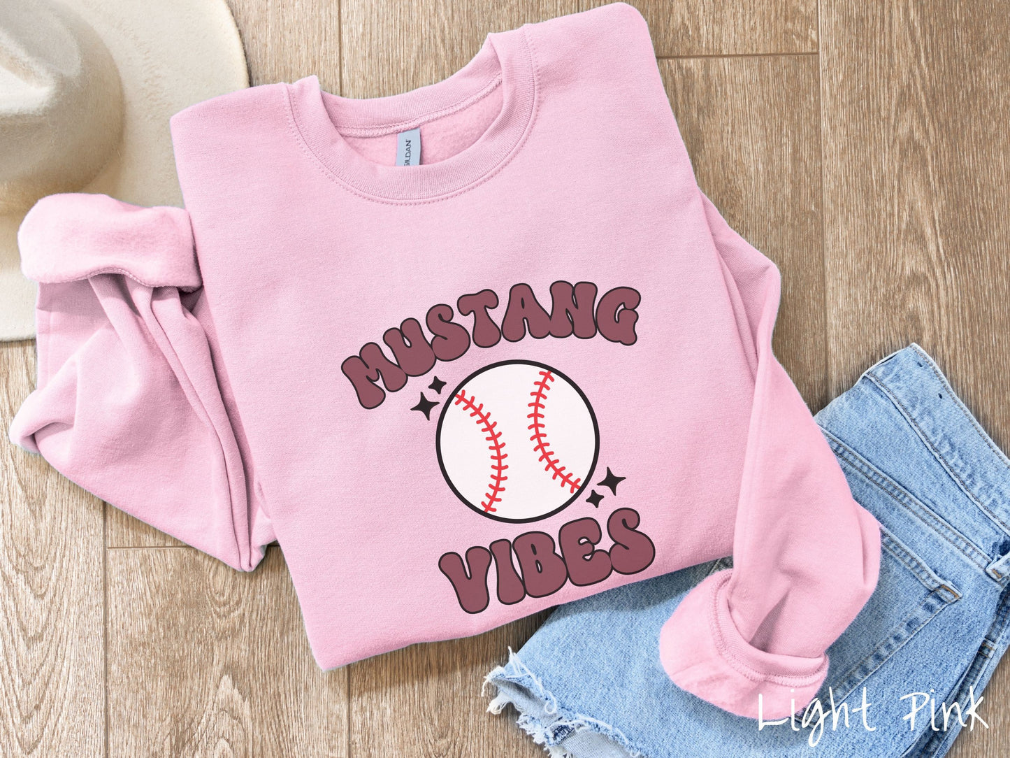 A woman wearing a cute light pink colored sweatshirt with purple text saying Mustang Vibes. There is a white baseball with red stitching between the text.