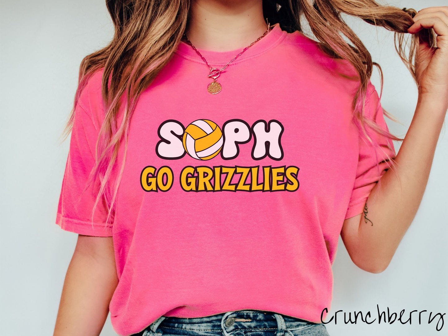 A woman wearing a cute crunchberry colored t-shirt with the word Sophin black and white font, the O in Soph is a yellow and white volleyball. Below Soph are the words in yellow and black font Go Grizzlies.