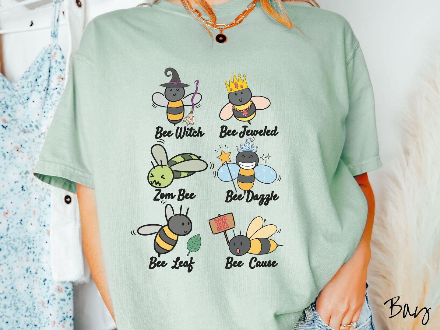 A woman wearing a cute, vintage bay colored comfy sweatshirt that has six puns that play on honey bees accompanied with bee drawings. The puns are Bee Witch, Bee Jeweled, Zom Bee, Bee Dazzle, Bee Leaf, and Bee Cause.