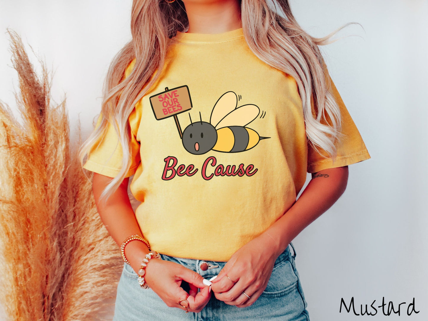 A woman wearing a cute, vintage mustard colored comfy sweatshirt with the text Bee Cause in red, cursive font. Above that is a black and yellow honey bee speaking with its mouth open and holding a sign that reads Save Our Bees in red font.
