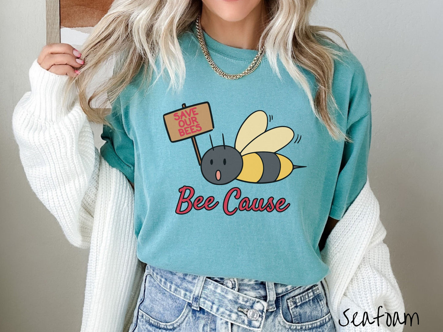 A woman wearing a cute, vintage seafoam colored comfy sweatshirt with the text Bee Cause in red, cursive font. Above that is a black and yellow honey bee speaking with its mouth open and holding a sign that reads Save Our Bees in red font.