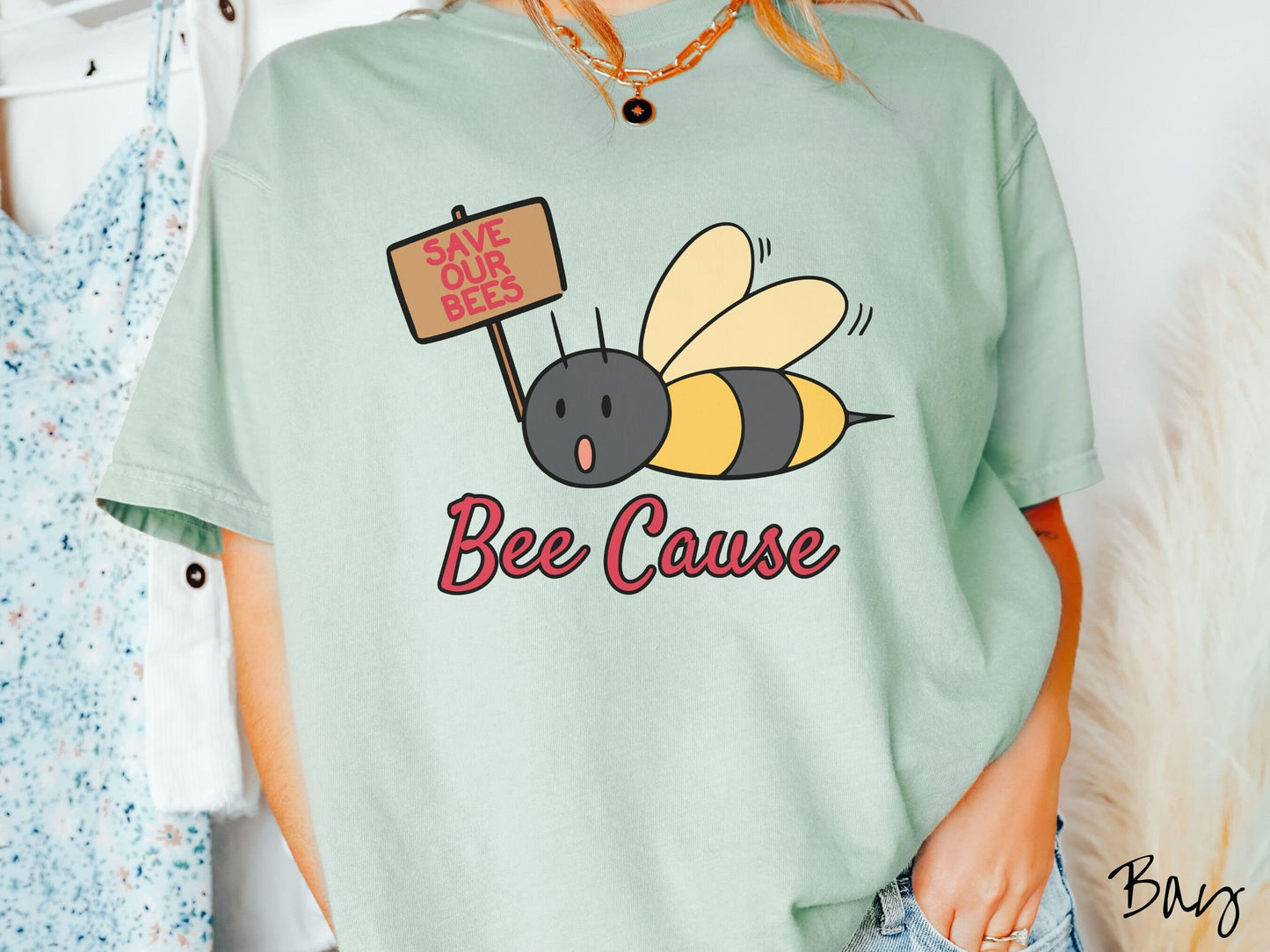 A woman wearing a cute, vintage bay colored comfy sweatshirt with the text Bee Cause in red, cursive font. Above that is a black and yellow honey bee speaking with its mouth open and holding a sign that reads Save Our Bees in red font.
