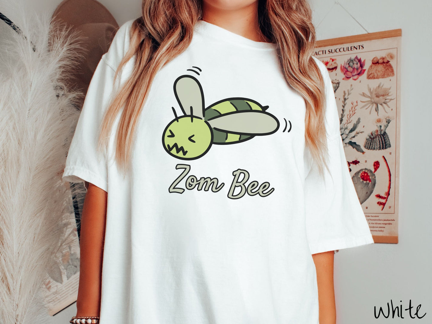 A woman wearing a cute, vintage white colored Comfort Colors t-shirt with the text Zom Bee in light green, cursive font. Above that is a light and dark green honey bee with a crazy face buzzing in the air.