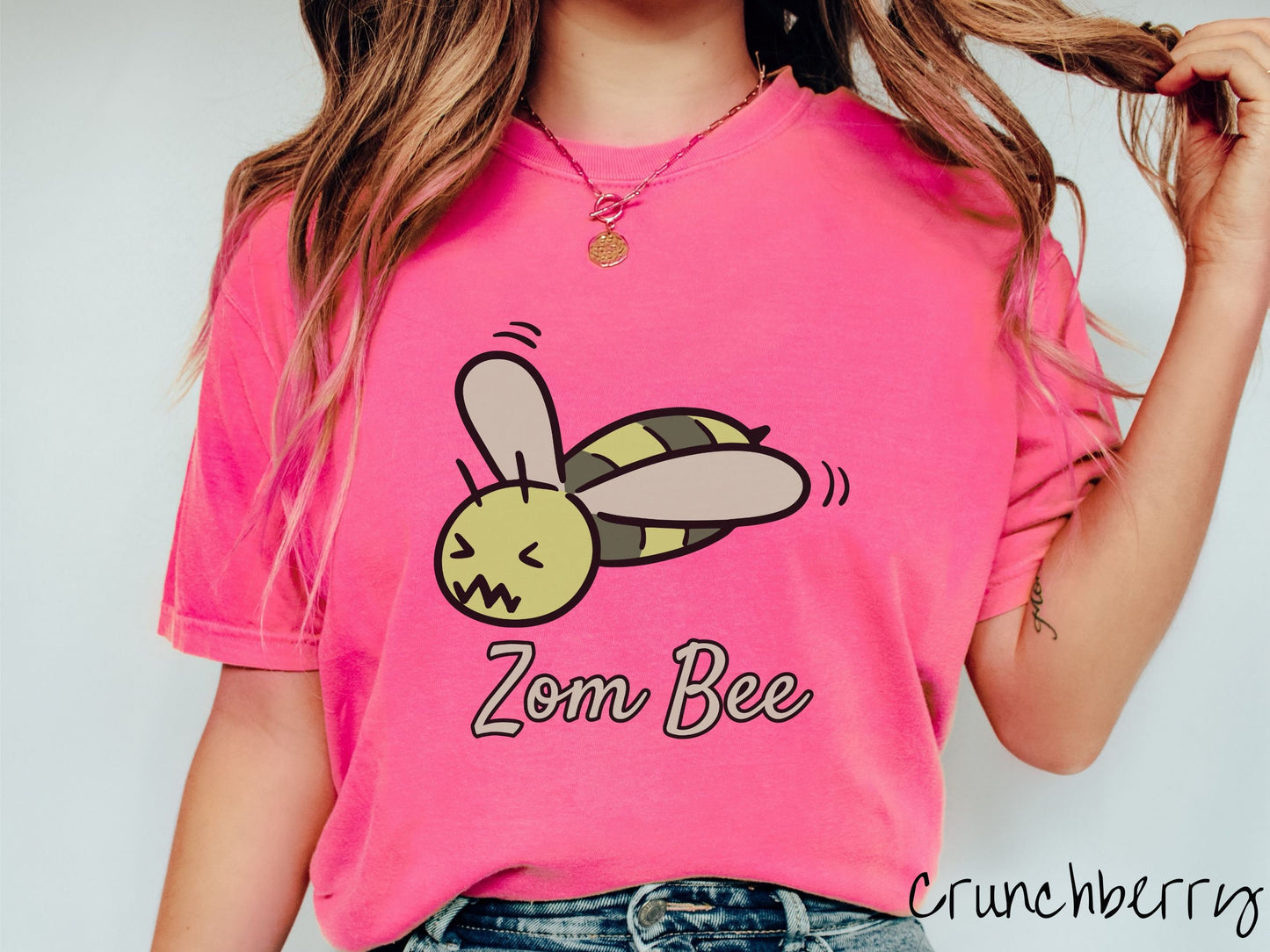 A woman wearing a cute, vintage crunchberry colored Comfort Colors t-shirt with the text Zom Bee in light green, cursive font. Above that is a light and dark green honey bee with a crazy face buzzing in the air.