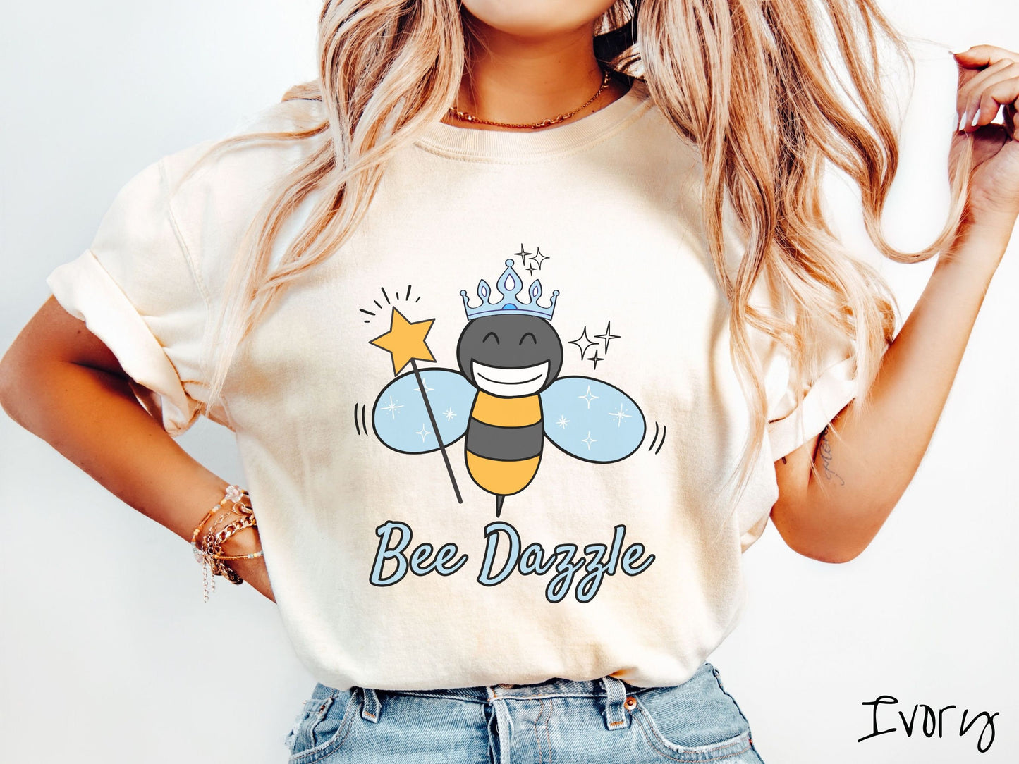 A woman wearing a cute, vintage ivory colored Comfort Colors t-shirt with text Bee Dazzle in light blue, cursive font. Above is a black and yellow bee with a wide smile and blue wings. It’s holding a star-tipped wand and wearing a light blue tiara.