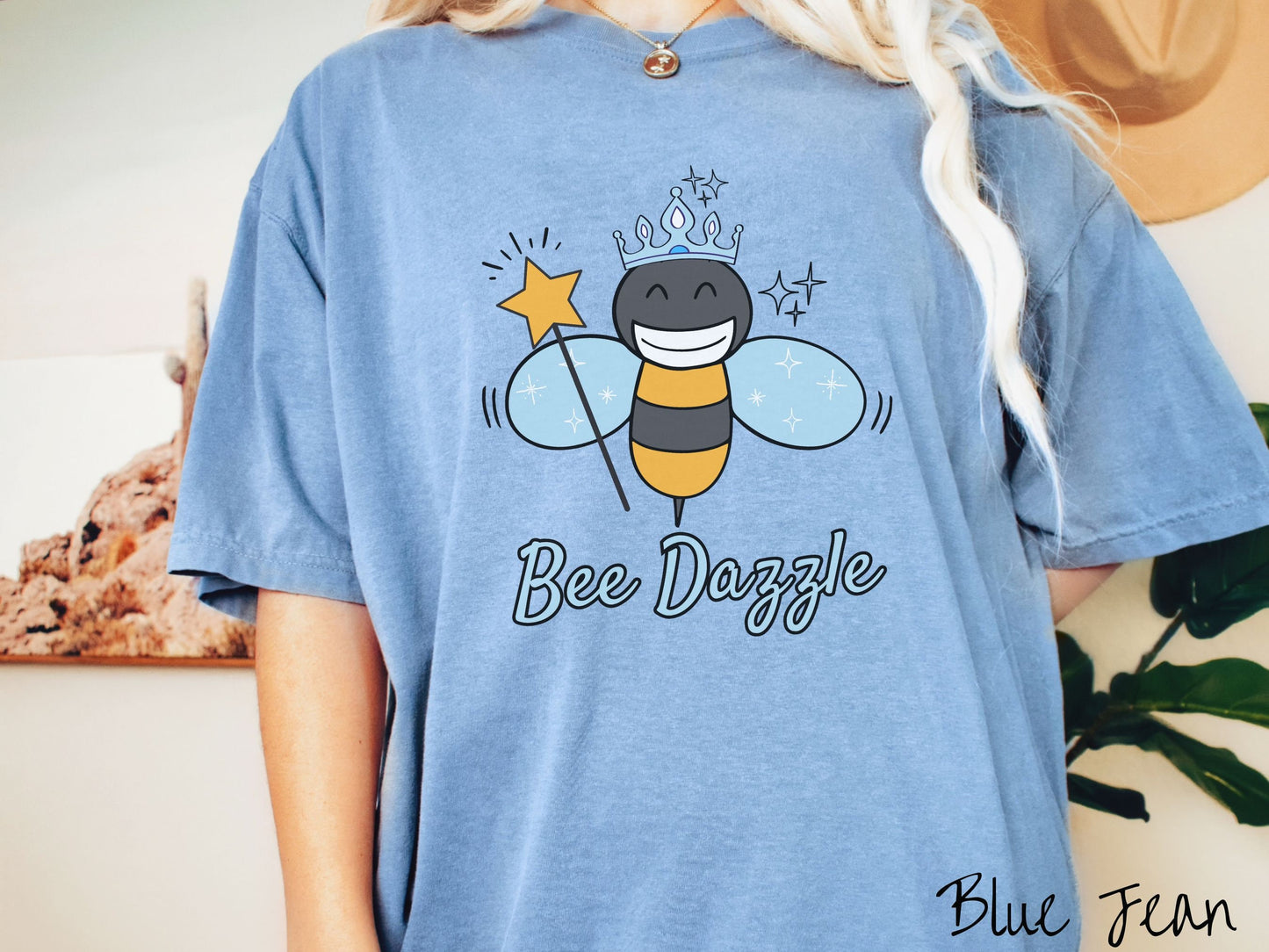 A woman wearing a cute, vintage blue jean colored Comfort Colors t-shirt with text Bee Dazzle in light blue, cursive font. Above is a black and yellow bee with a wide smile and blue wings. It’s holding a star-tipped wand and wearing a blue tiara.