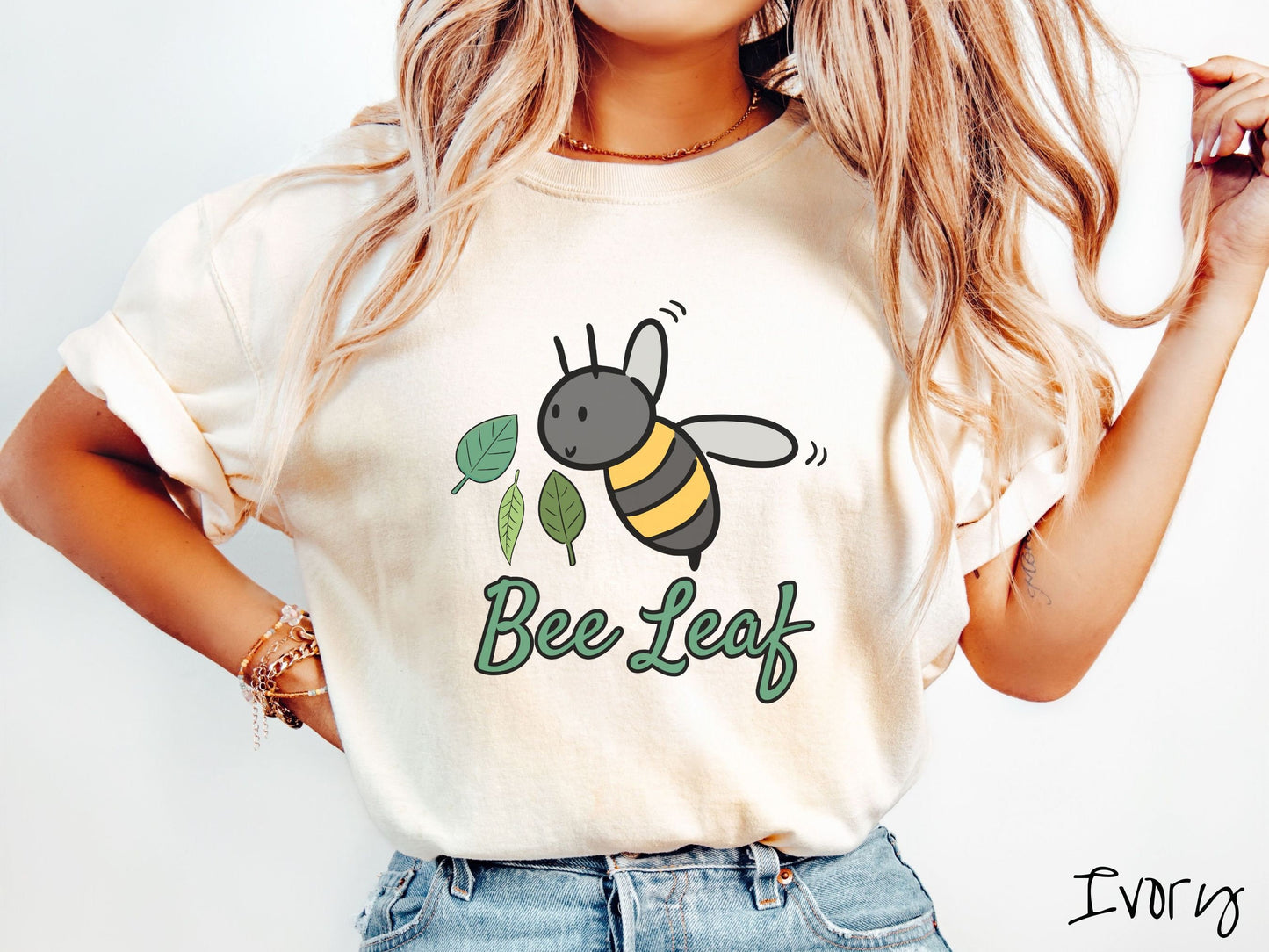 A woman wearing a cute, vintage ivory colored Comfort Colors t-shirt with the text Bee Leaf in green, cursive font. Above that is a black and yellow honey bee buzzing through the air with green leaves floating next to it.