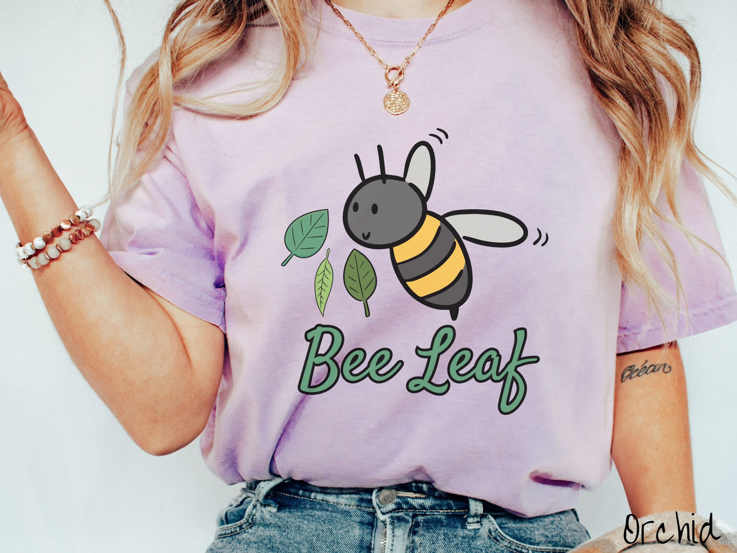 A woman wearing a cute, vintage orchid colored Comfort Colors t-shirt with the text Bee Leaf in green, cursive font. Above that is a black and yellow honey bee buzzing through the air with green leaves floating next to it.