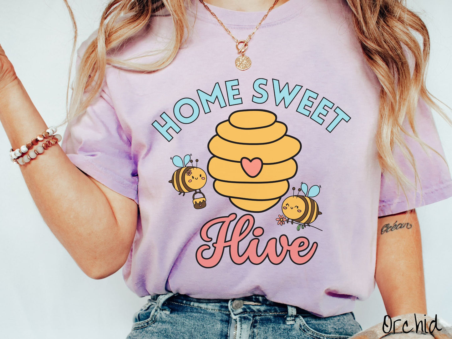 A woman wearing a cute, vintage orchid colored Comfort Colors t-shirt with the text Home Sweet Hive in cyan and red font. In between the words are a yellow bee hive with a red heart on it and smiling honey bees flying around holding flowers.