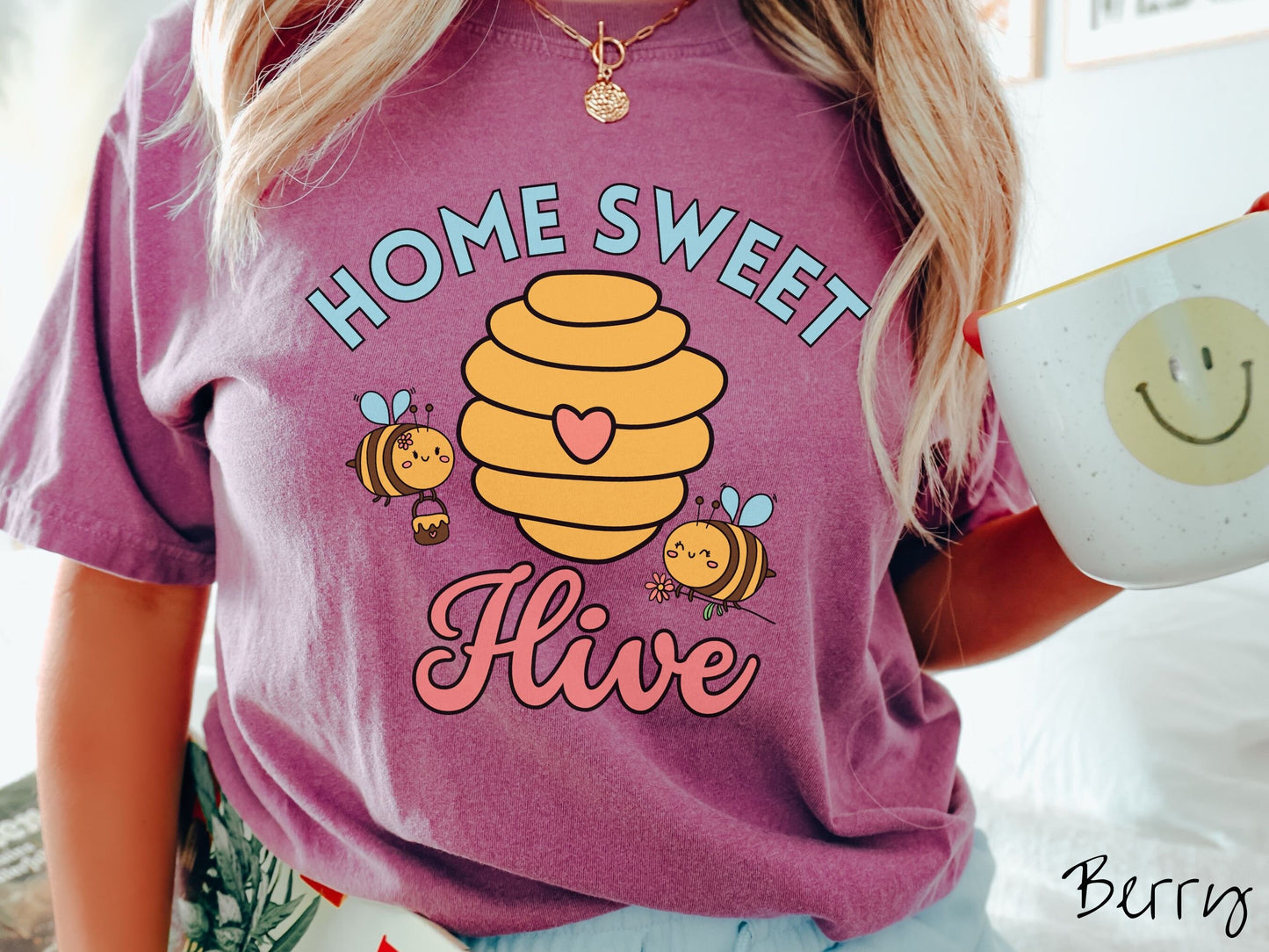 A woman wearing a cute, vintage berry colored Comfort Colors t-shirt with the text Home Sweet Hive in cyan and red font. In between the words are a yellow bee hive with a red heart on it and smiling honey bees flying around holding flowers.