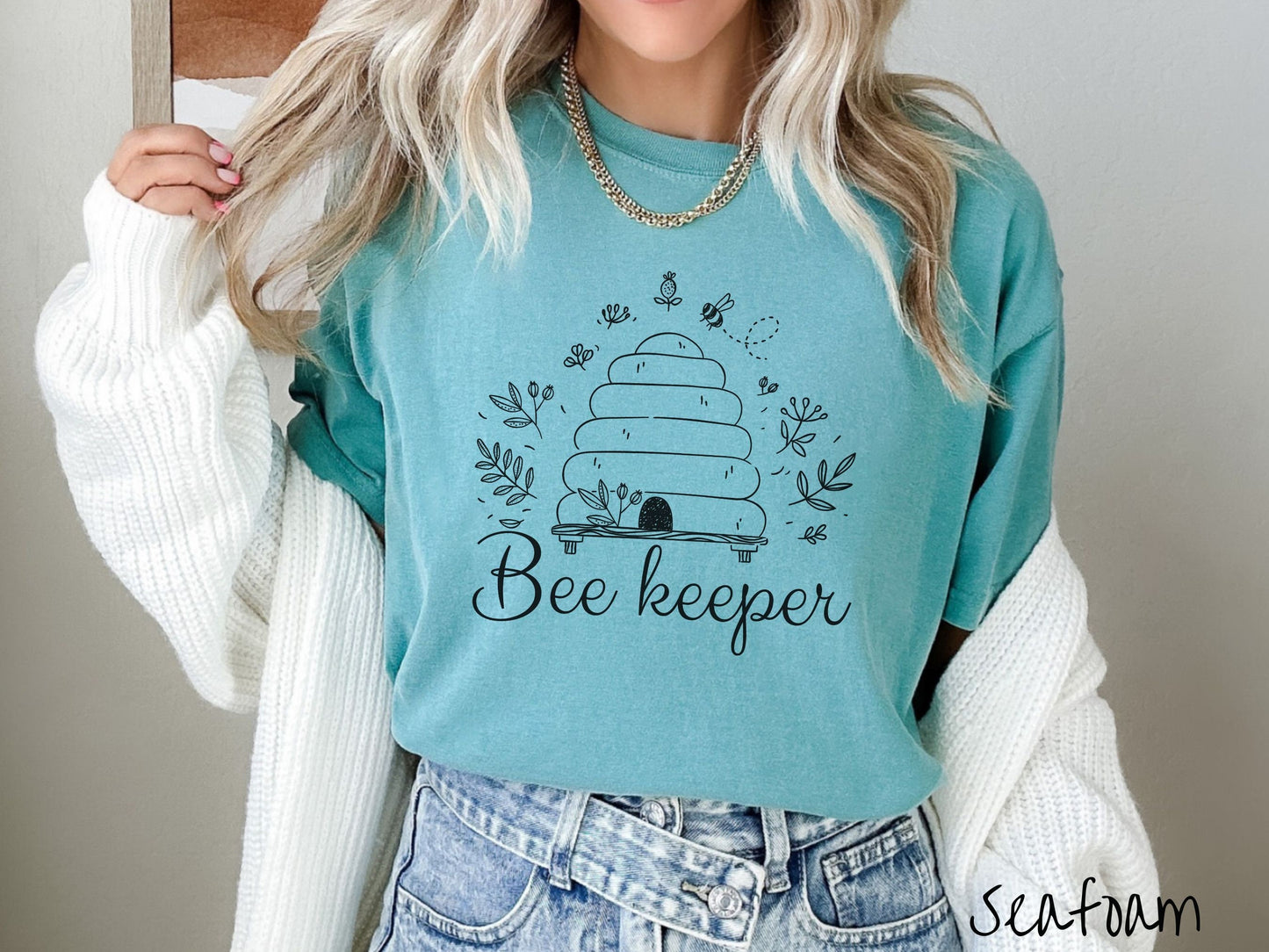 A woman wearing a cute, vintage seafoam colored Comfort Colors t-shirt with the text Beekeeper across the front in black, cursive font. Above this is a black-outlined bee hive with bees buzzing around it and there are leaves in the air.