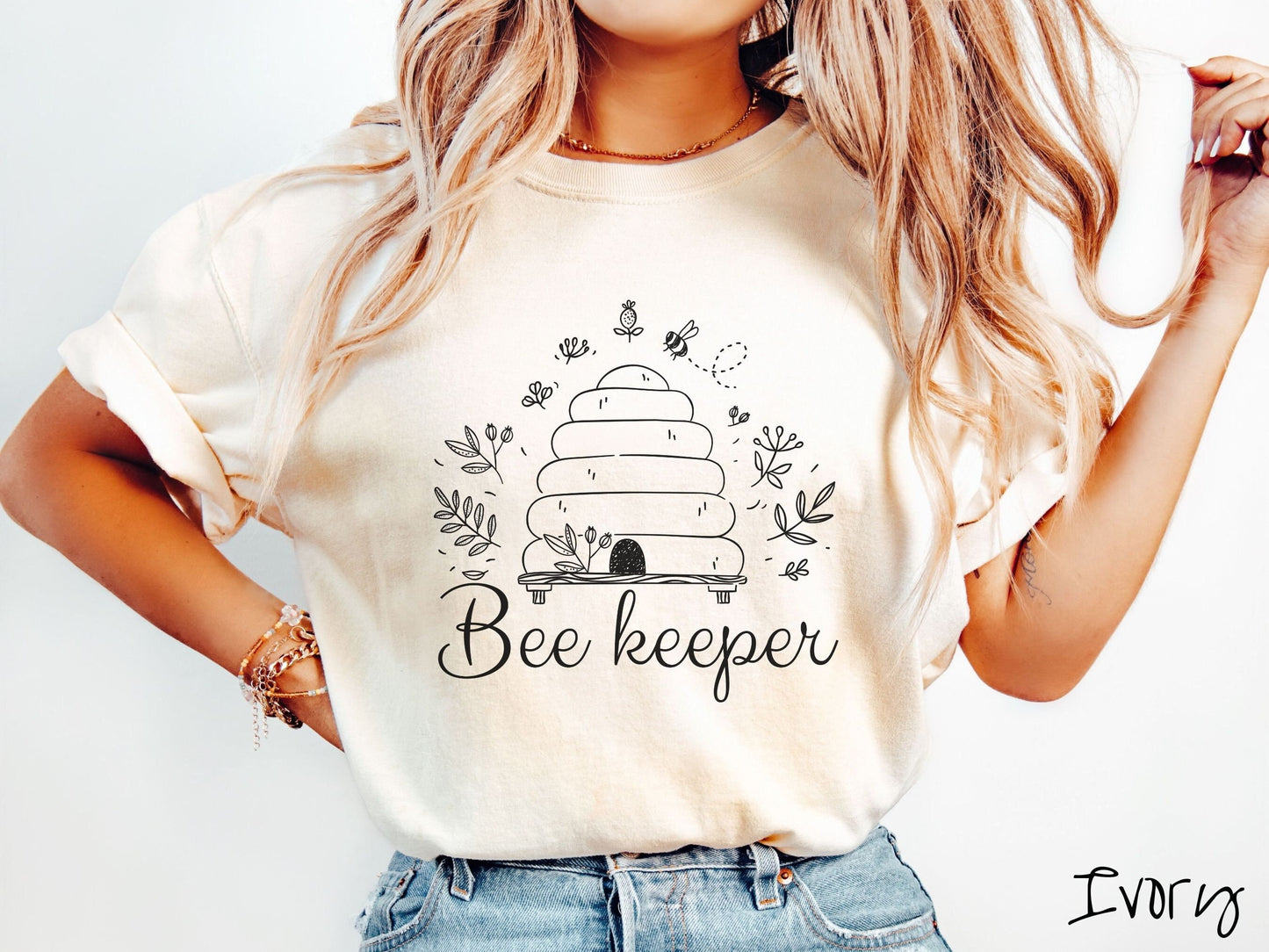 A woman wearing a cute, vintage ivory colored Comfort Colors t-shirt with the text Beekeeper across the front in black, cursive font. Above this is a black-outlined bee hive with bees buzzing around it and there are leaves in the air.