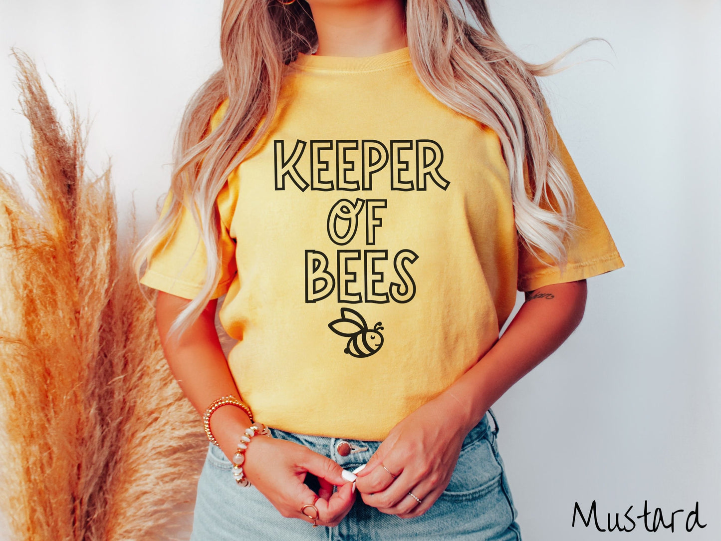 A woman wearing a cute, vintage mustard colored Comfort Colors t-shirt with the text Keeper of Bees in black uppercase font. Below the text is a black-outlined smiling bee looking up.