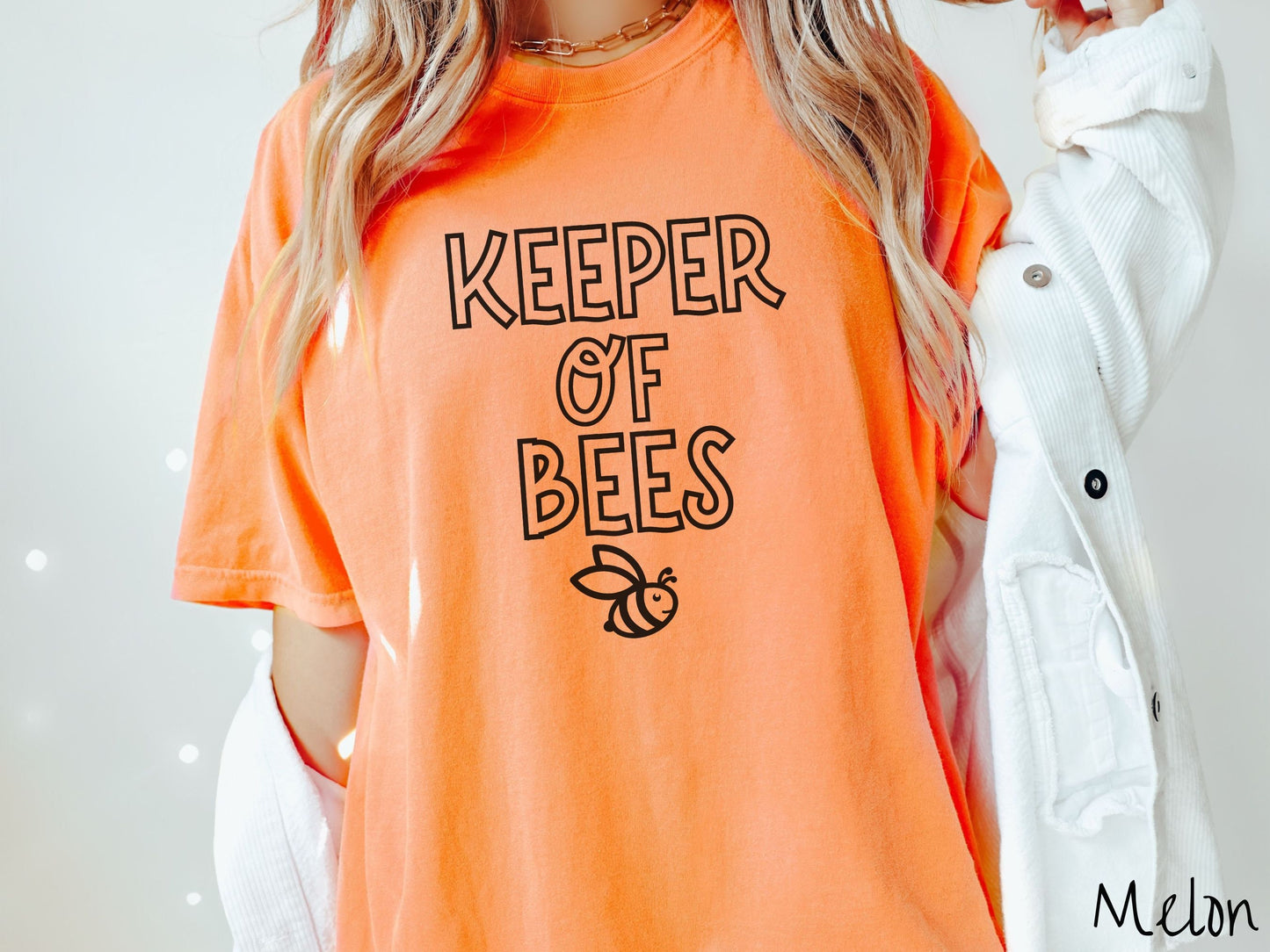A woman wearing a cute, vintage melon colored Comfort Colors t-shirt with the text Keeper of Bees in black uppercase font. Below the text is a black-outlined smiling bee looking up.