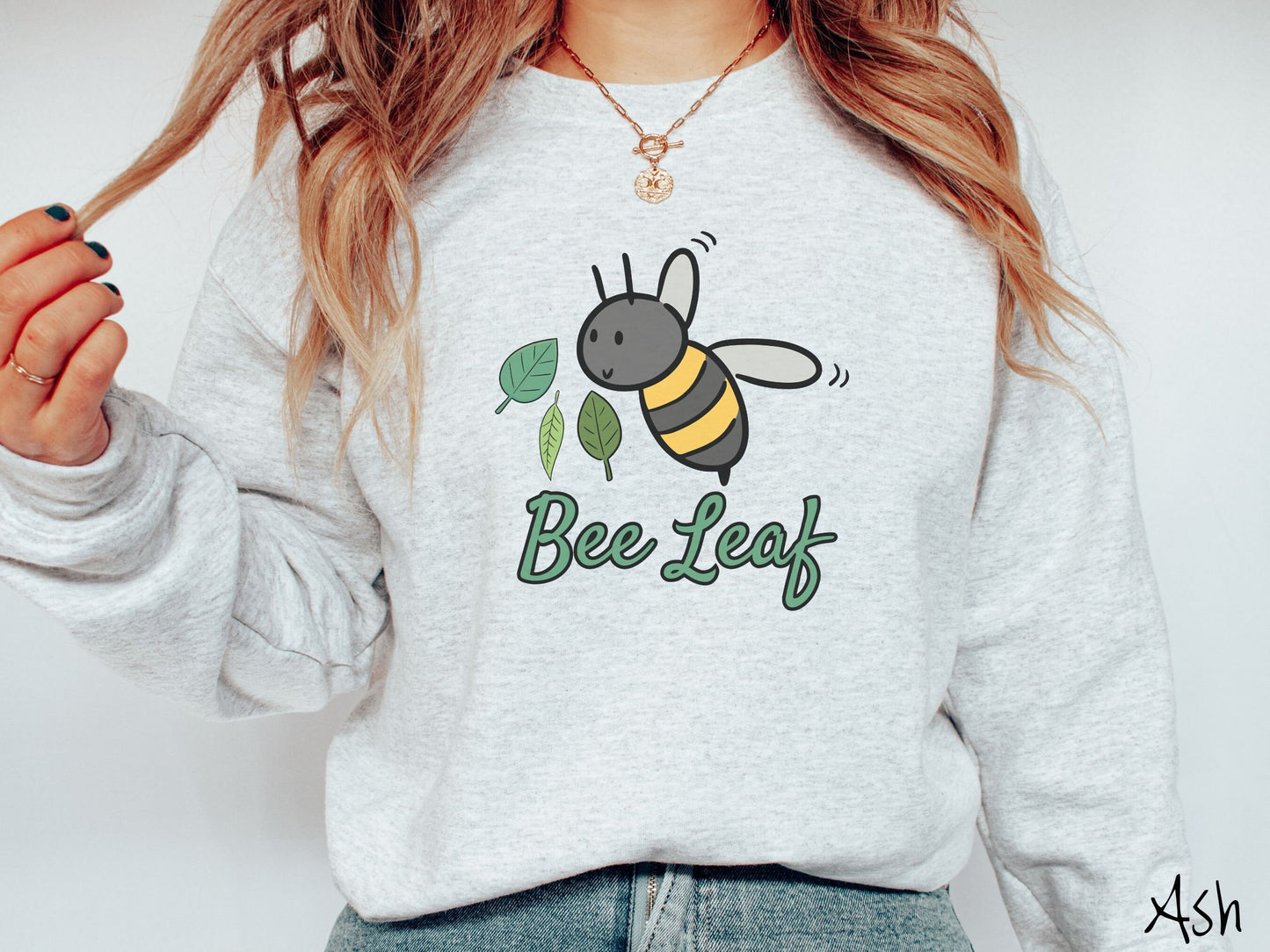 A woman wearing a cute, vintage ash colored comfy sweatshirt with the text Bee Leaf in green, cursive font. Above that is a black and yellow honey bee buzzing through the air with green leaves floating next to it.