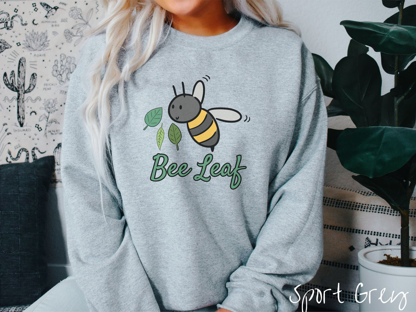 A woman wearing a cute, vintage sport grey colored comfy sweatshirt with the text Bee Leaf in green, cursive font. Above that is a black and yellow honey bee buzzing through the air with green leaves floating next to it.