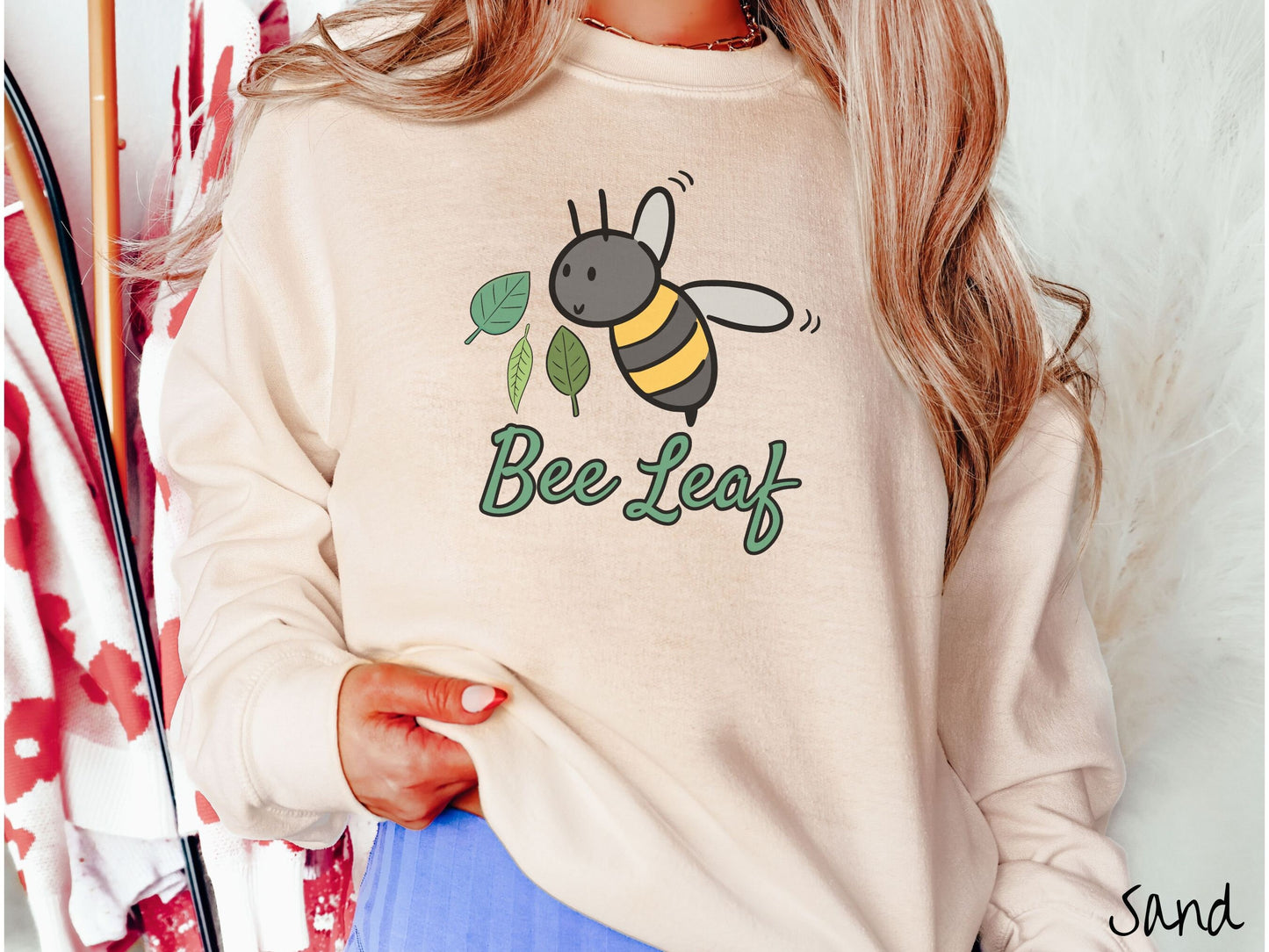 A woman wearing a cute, vintage sand colored comfy sweatshirt with the text Bee Leaf in green, cursive font. Above that is a black and yellow honey bee buzzing through the air with green leaves floating next to it.