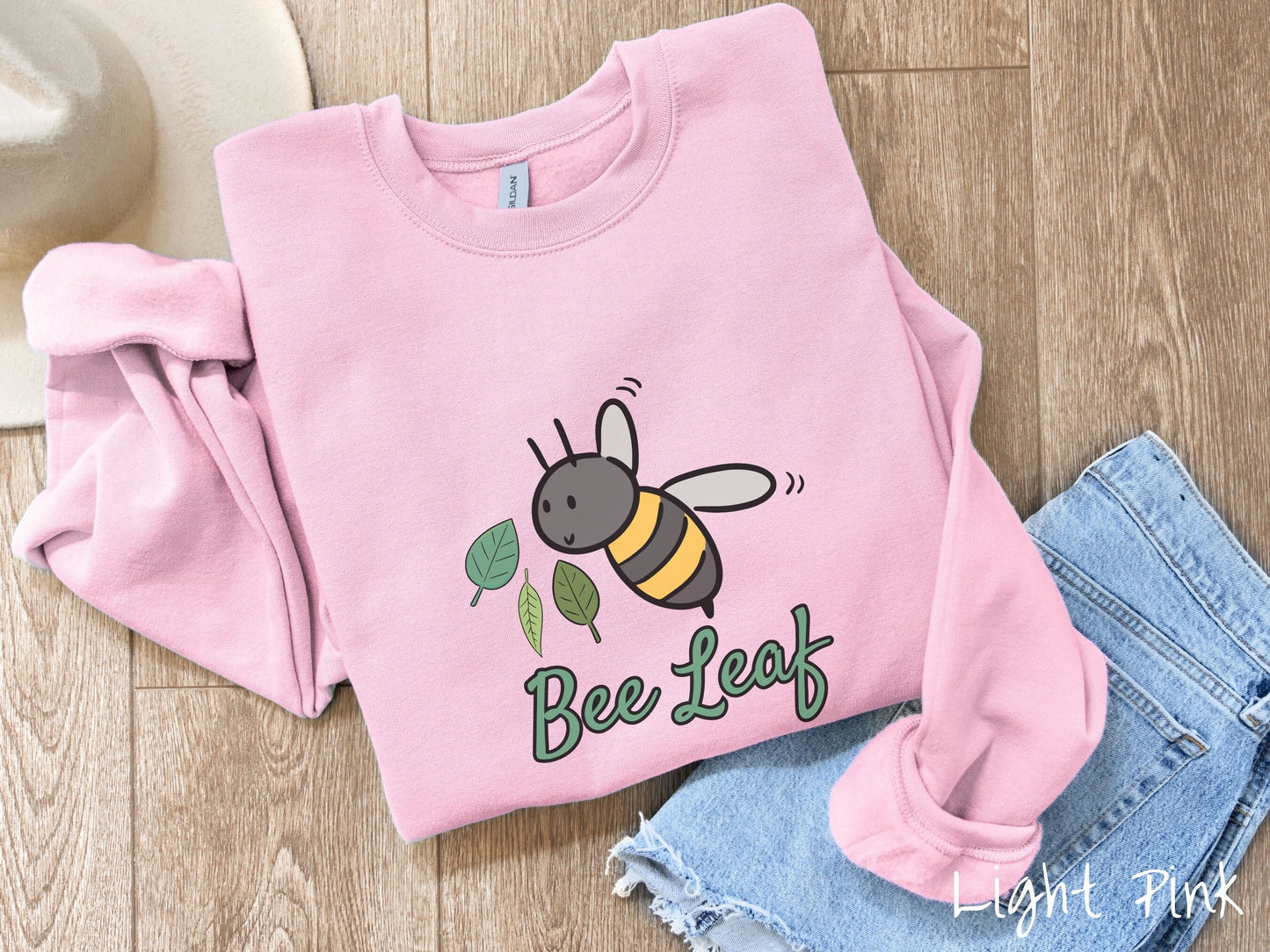 A cute, vintage light pink colored comfy sweatshirt with the text Bee Leaf in green, cursive font. Above that is a black and yellow honey bee buzzing through the air with green leaves floating next to it.