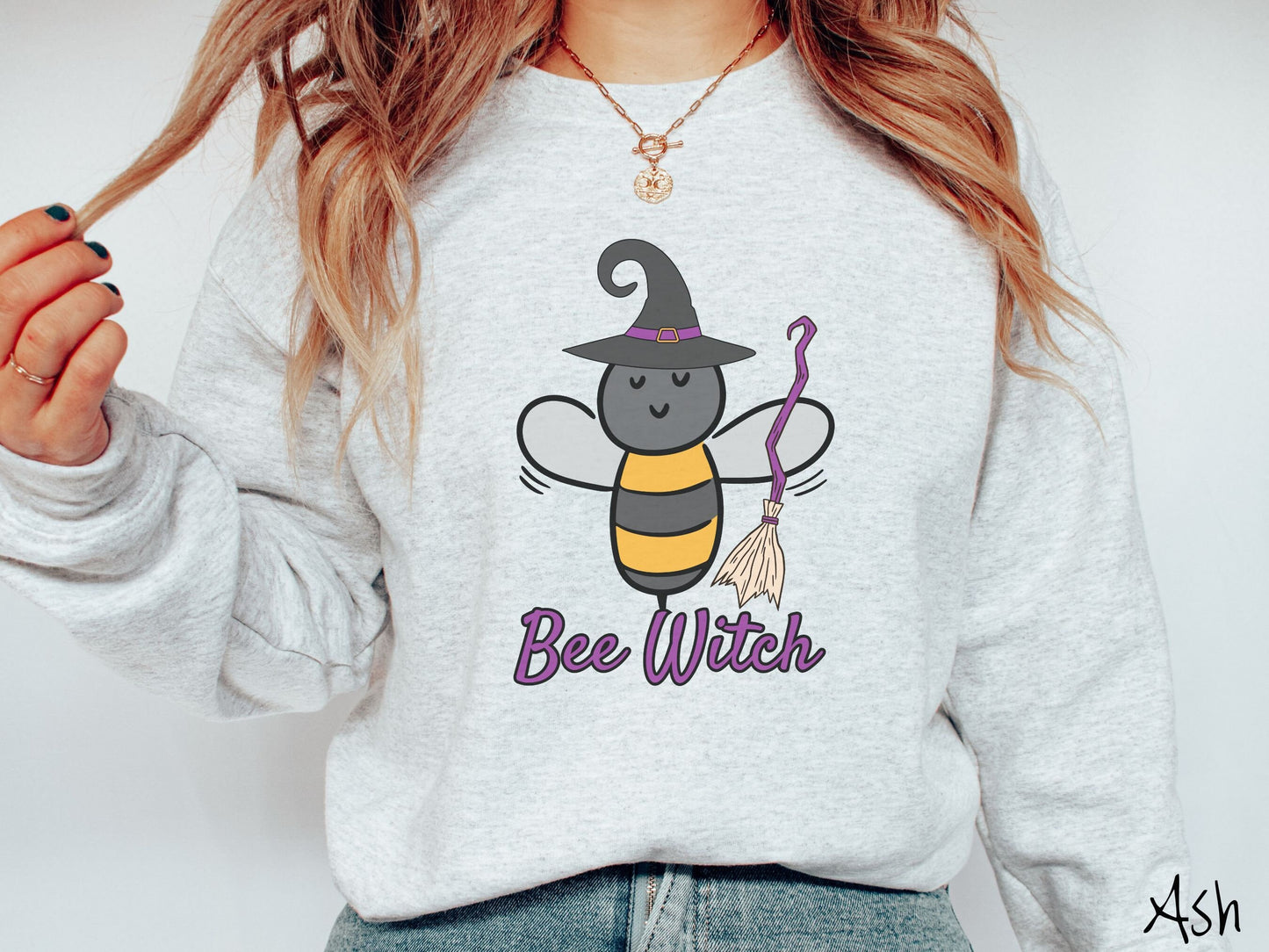 A woman wearing a cute, vintage ash colored comfy sweatshirt with the text Bee Witch in purple, cursive font. Above that is a black and yellow honey bee holding a purple, straw broom and wearing a black witch hat with a purple hat band.