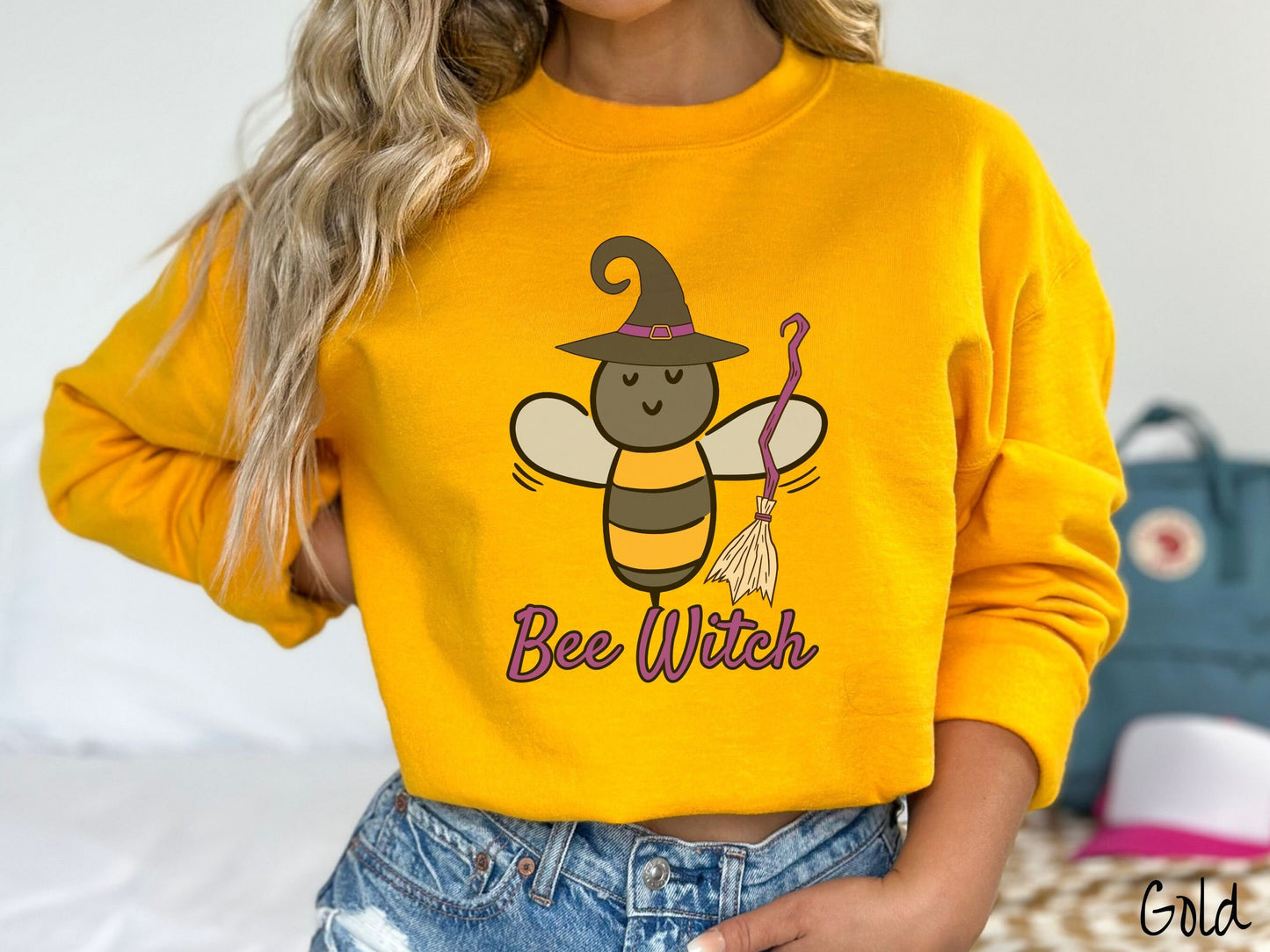 A woman wearing a cute, vintage gold colored comfy sweatshirt with the text Bee Witch in purple, cursive font. Above that is a black and yellow honey bee holding a purple, straw broom and wearing a black witch hat with a purple hat band.v