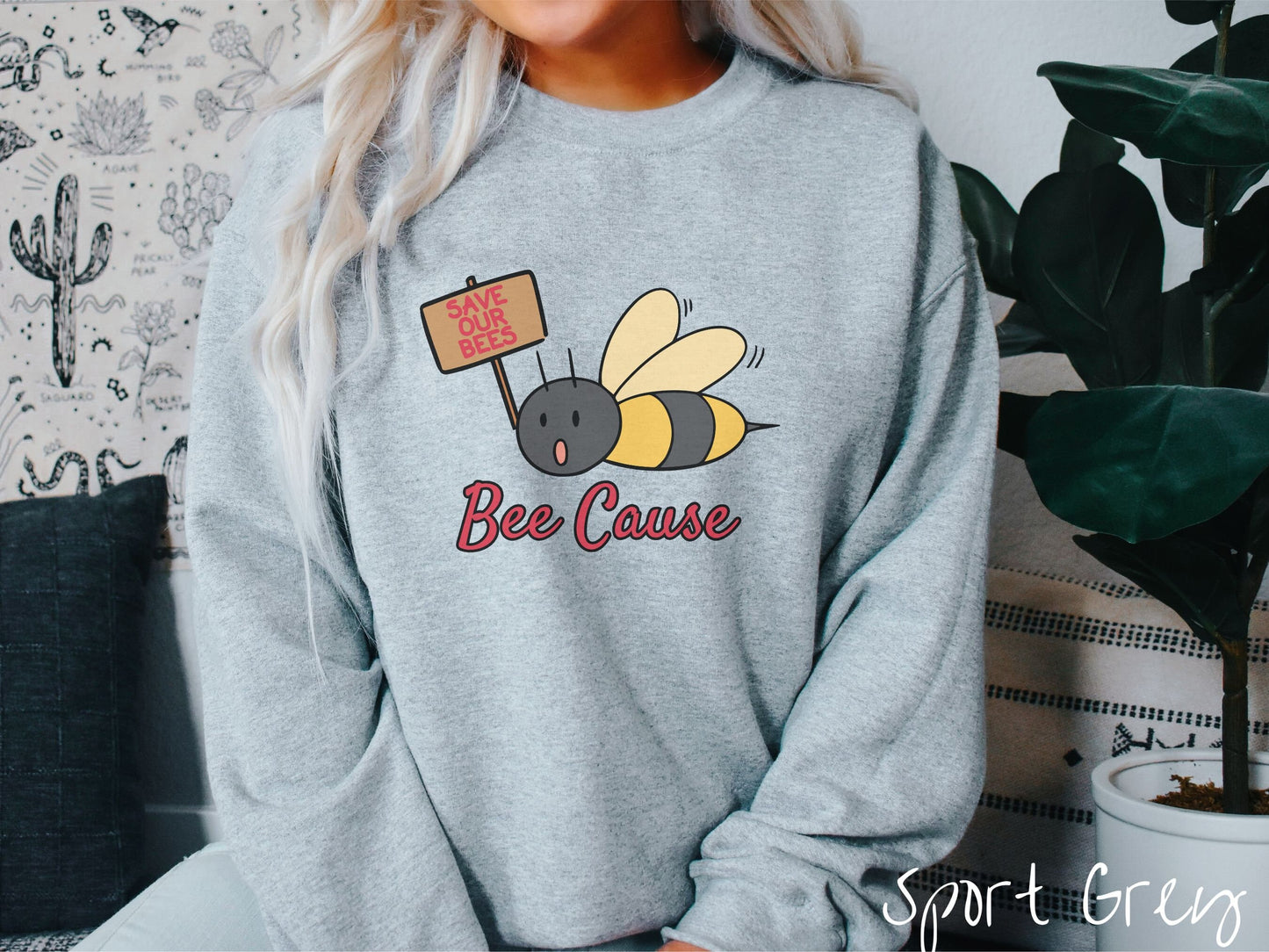 A woman wearing a cute, vintage sport grey colored comfy sweatshirt with the text Bee Cause in red, cursive font. Above that is a black and yellow honey bee speaking with its mouth open and holding a sign that reads Save Our Bees in red font.