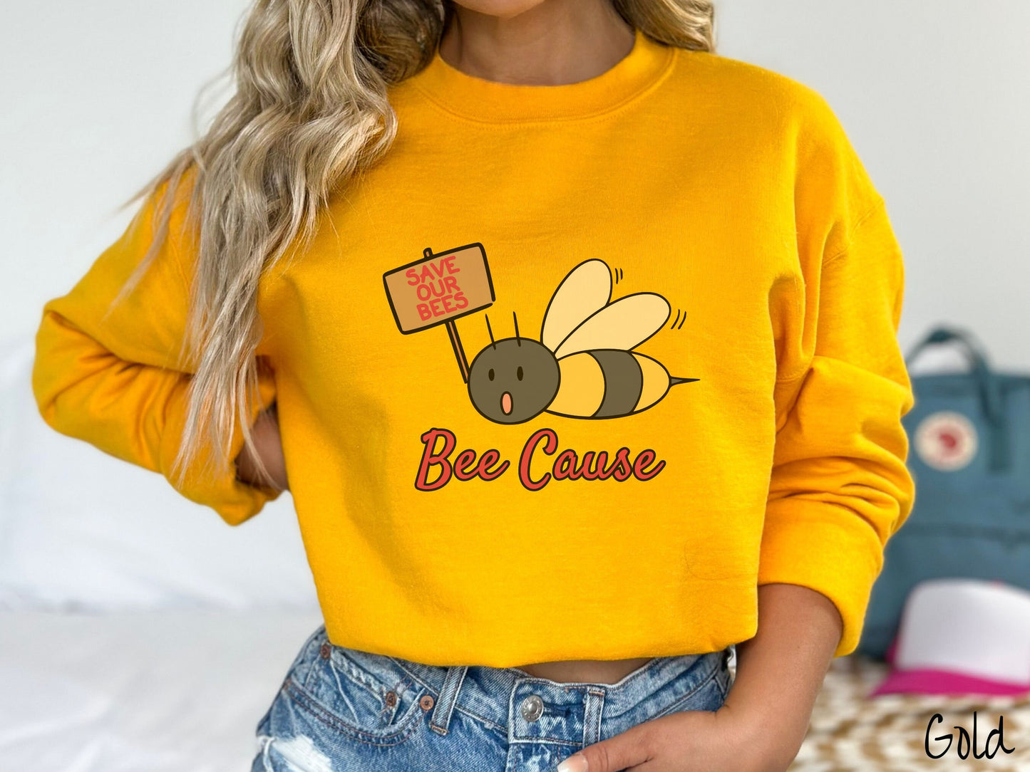 A woman wearing a cute, vintage gold colored comfy sweatshirt with the text Bee Cause in red, cursive font. Above that is a black and yellow honey bee speaking with its mouth open and holding a sign that reads Save Our Bees in red font.