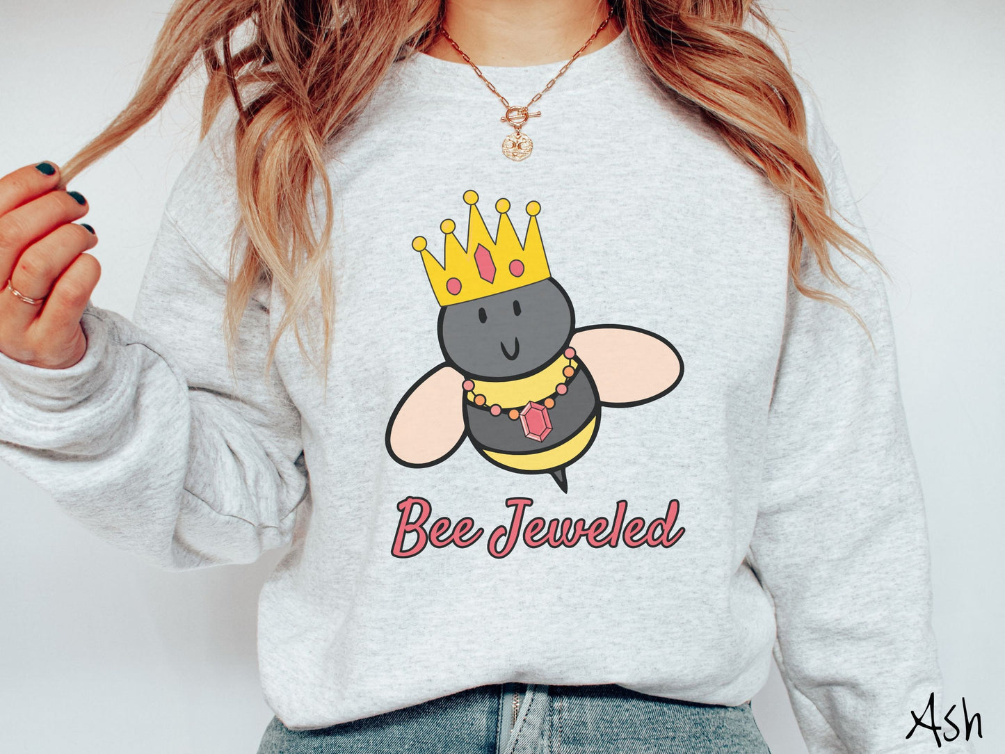A woman wearing a cute, vintage ash colored comfy sweatshirt with the text Bee Jeweled in pink, cursive font. Above that is a black and yellow honey bee wearing a red, bejeweled golden crown, and a golden necklace with a large, red jewel.
