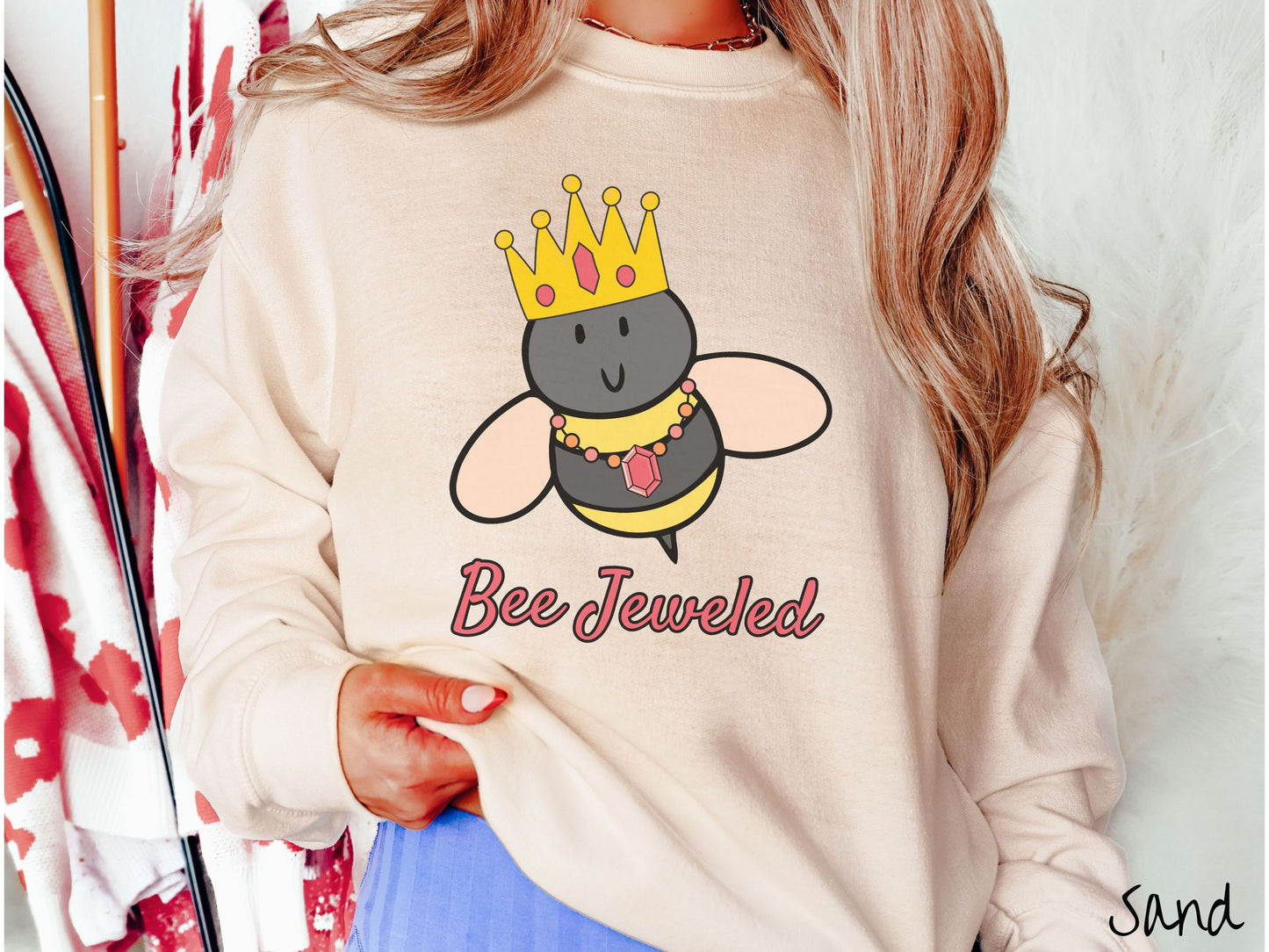 A woman wearing a cute, vintage sand colored comfy sweatshirt with the text Bee Jeweled in pink, cursive font. Above that is a black and yellow honey bee wearing a red, bejeweled golden crown, and a golden necklace with a large, red jewel.