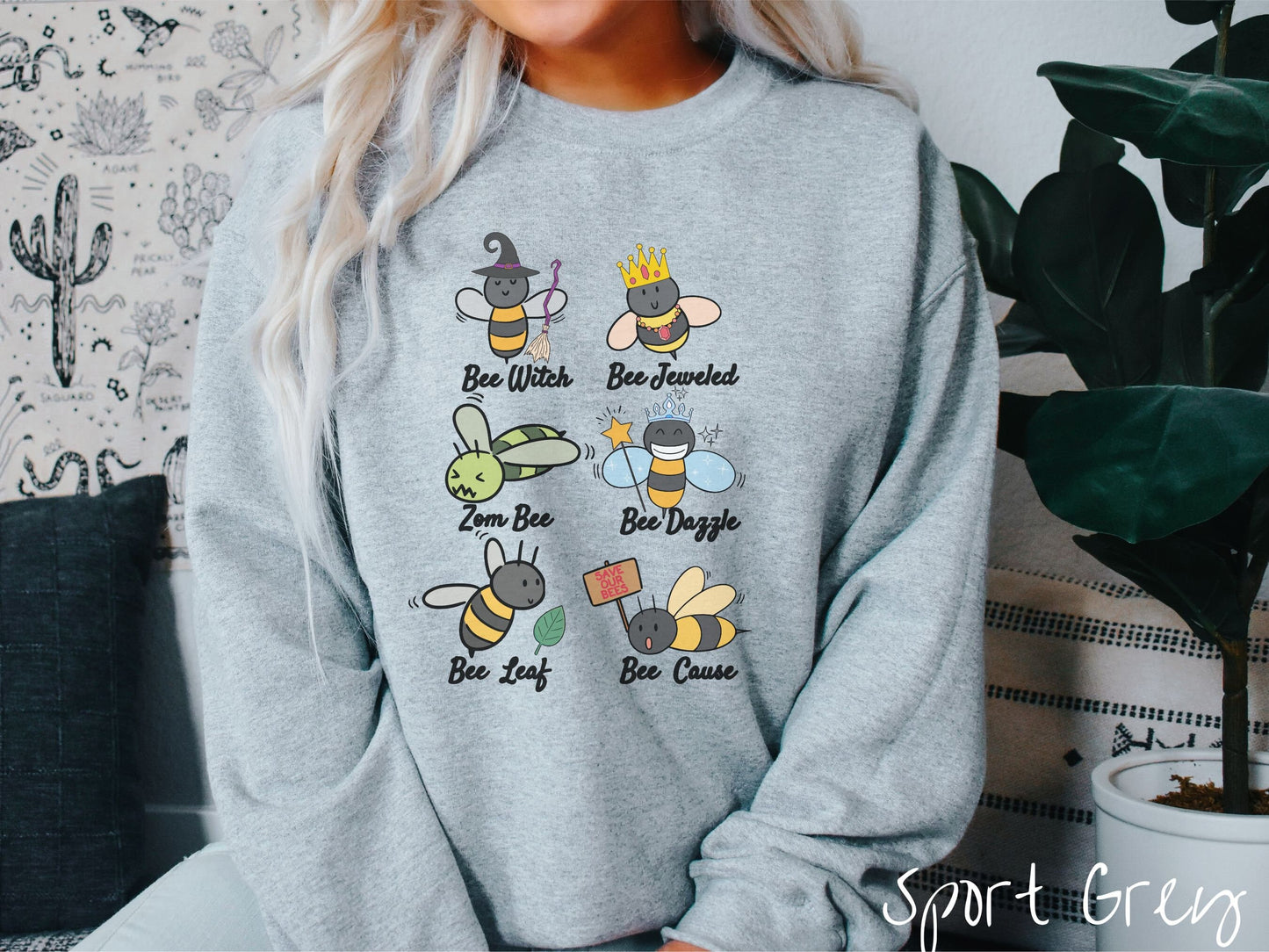 A woman wearing a cute, vintage sport grey colored comfy sweatshirt that has six puns that play on honey bees accompanied with bee drawings. The puns are Bee Witch, Bee Jeweled, Zom Bee, Bee Dazzle, Bee Leaf, and Bee Cause.