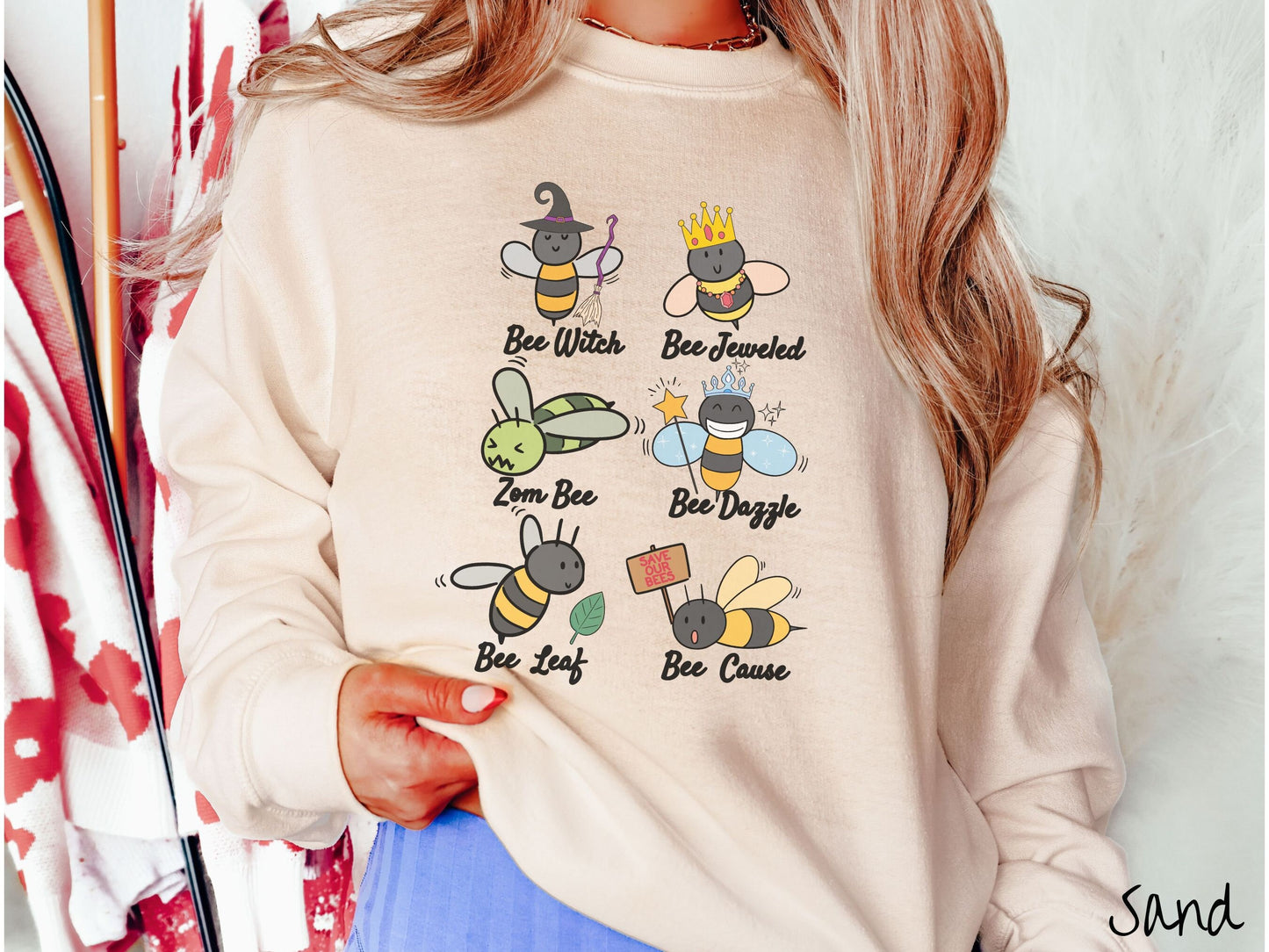 A woman wearing a cute, vintage sand colored comfy sweatshirt that has six puns that play on honey bees accompanied with bee drawings. The puns are Bee Witch, Bee Jeweled, Zom Bee, Bee Dazzle, Bee Leaf, and Bee Cause.